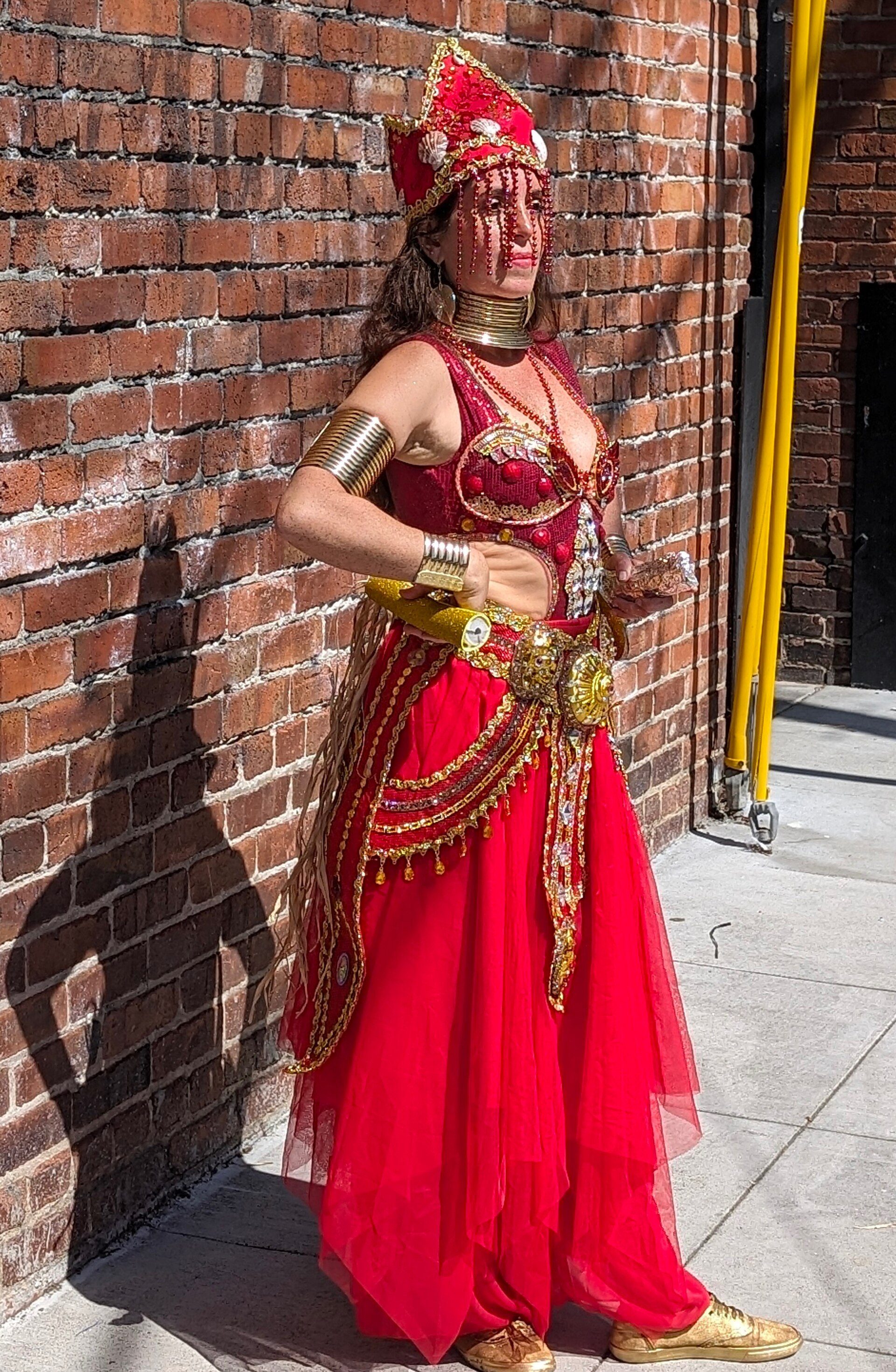 A person in a vibrant red and gold costume stands against a brick wall.