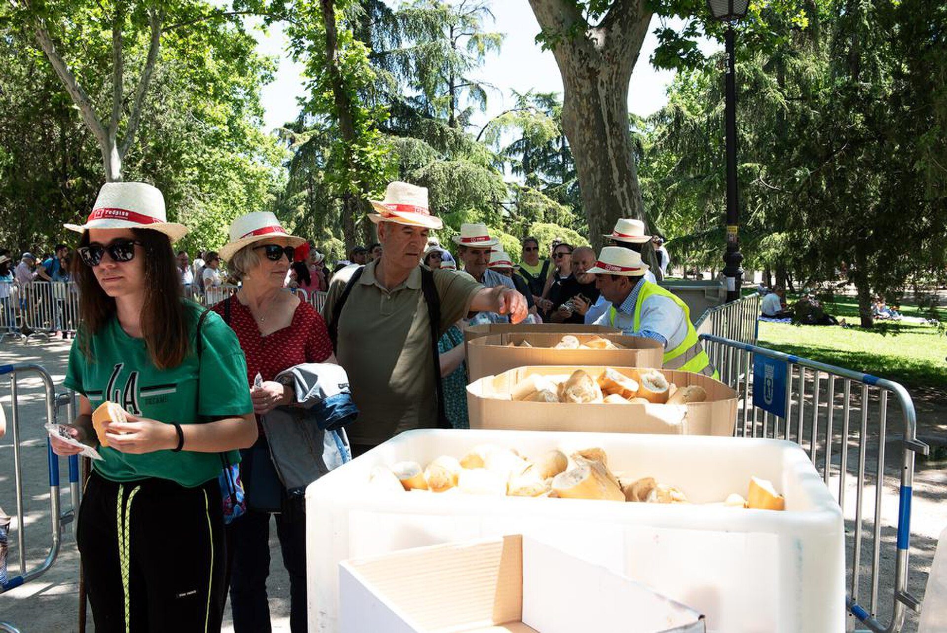 Alt text: Outdoor scene with people in hats, food boxes, and trees in a park setting.