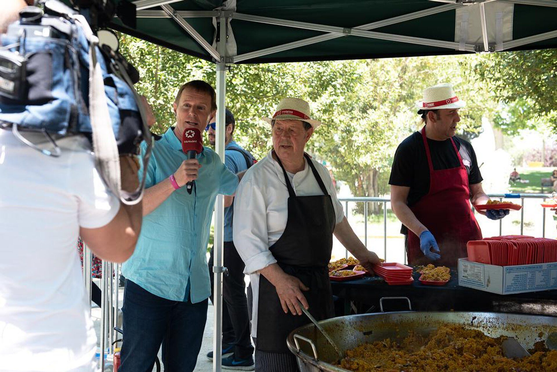 A man in a chef's hat is cooking at an outdoor event, with a reporter interviewing him.