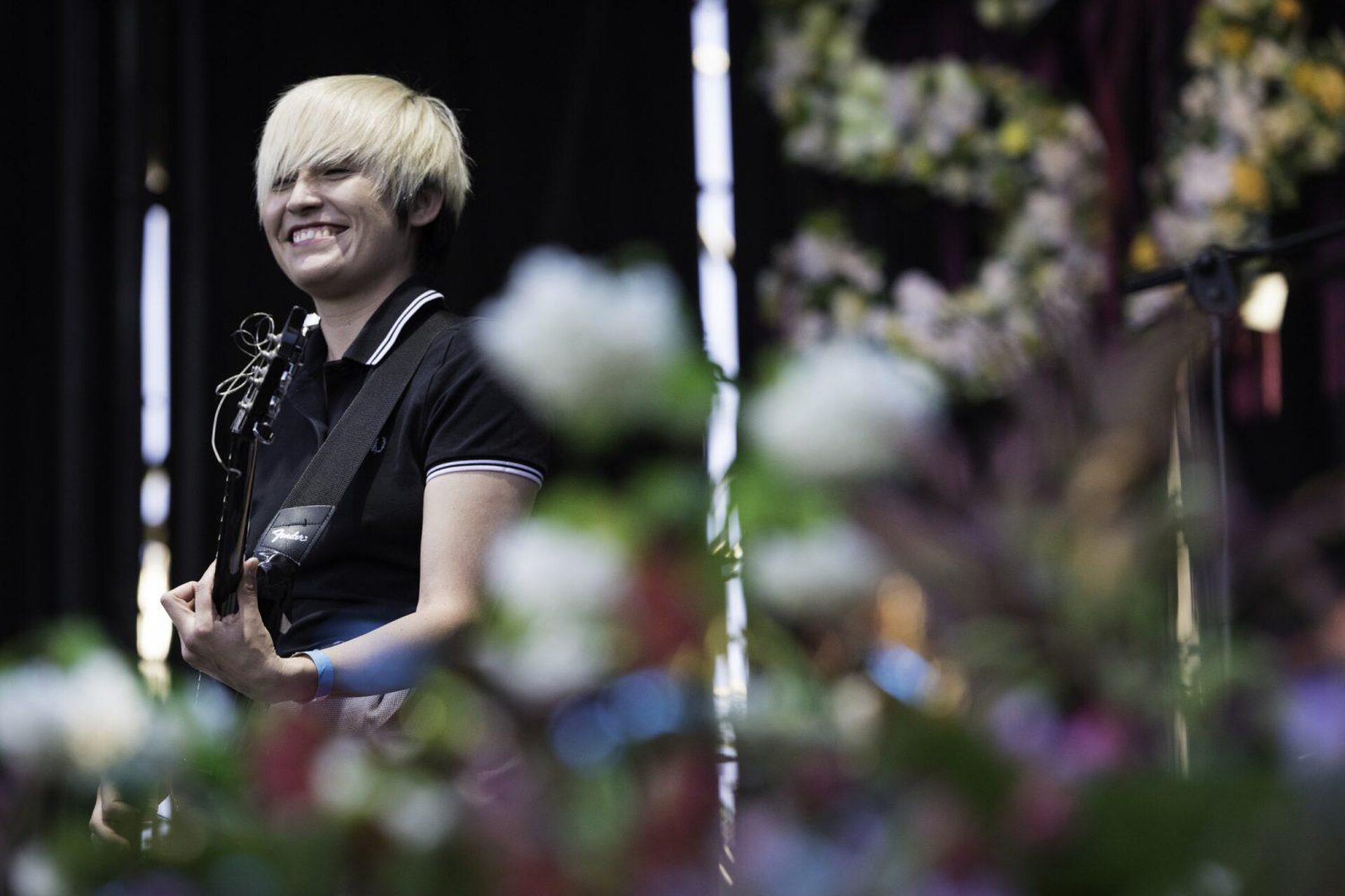 A person with short blonde hair smiling, holding a guitar on stage with floral decorations.