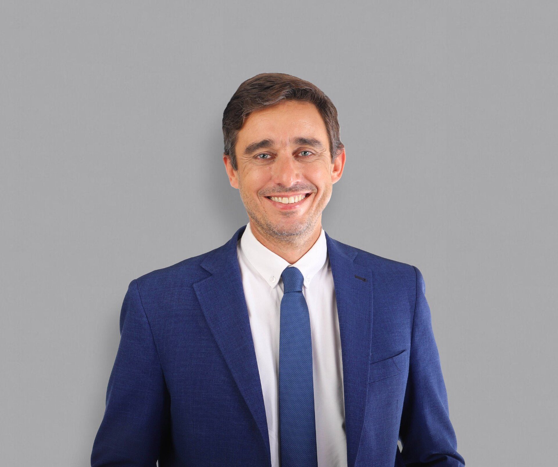 A man in a blue suit and tie stands against a gray background.