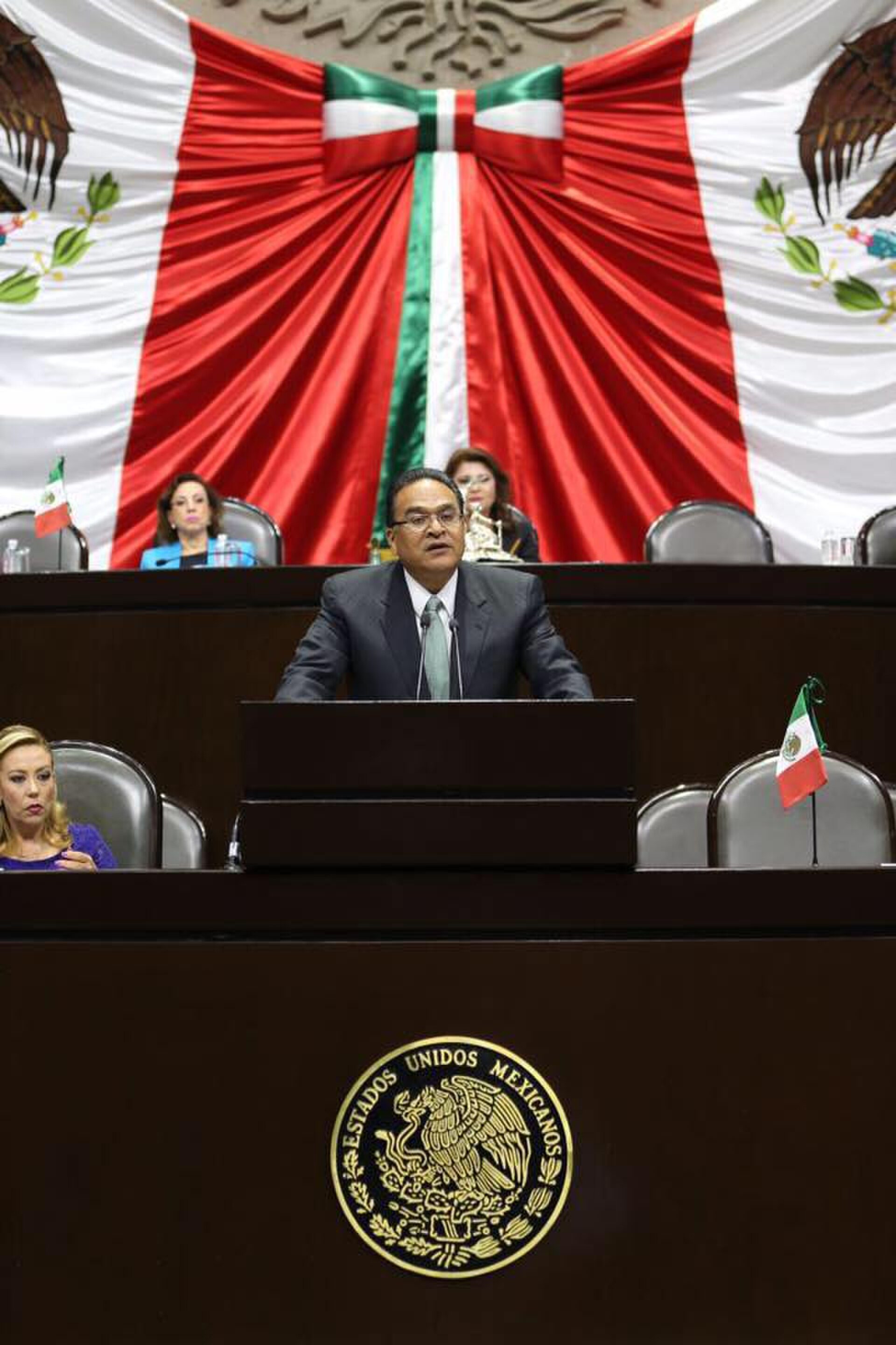 A formal legislative chamber with a podium, flags, and a large Mexican flag backdrop.