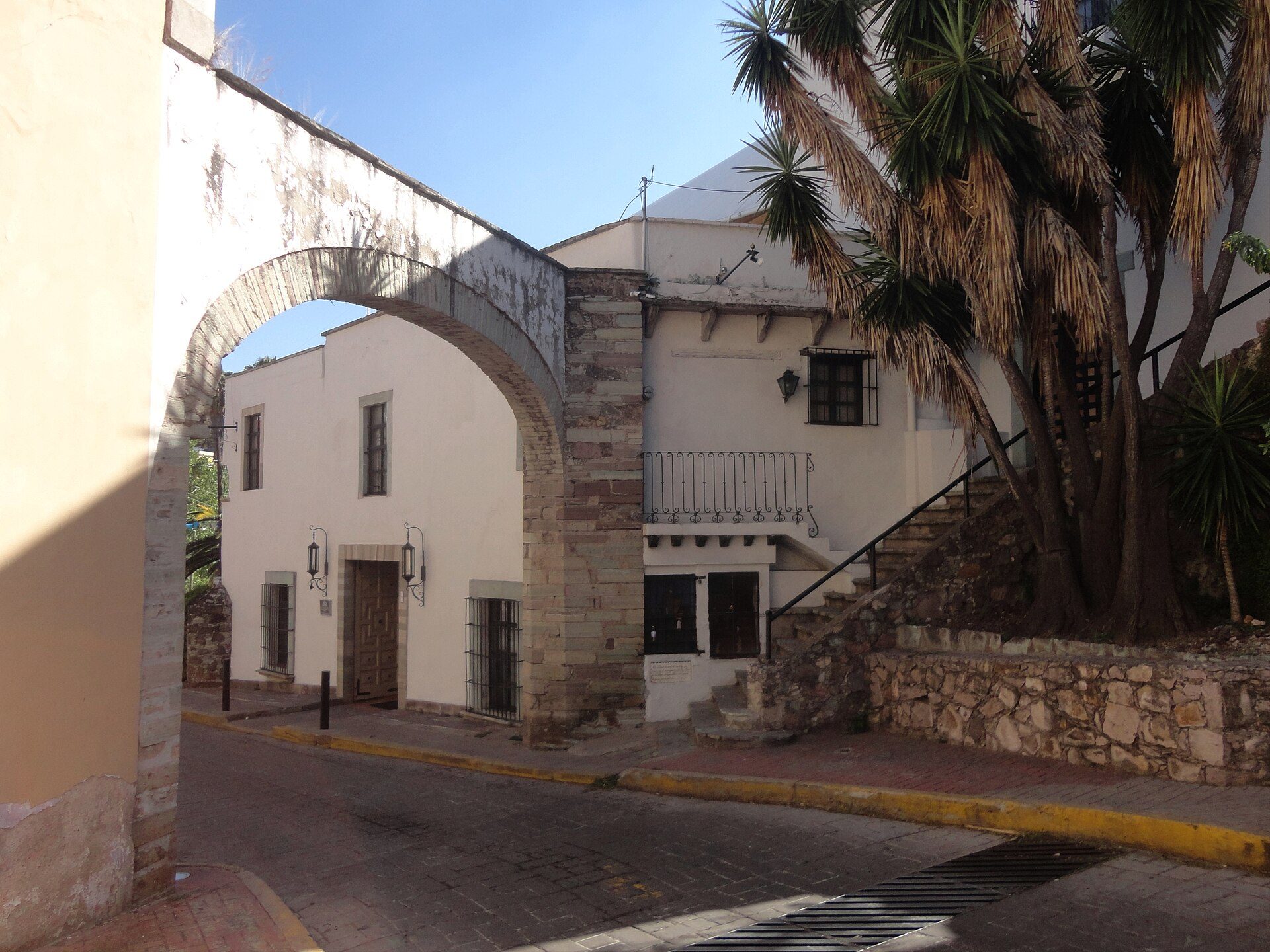 Traditional white-walled building with arched entrance, stone staircase, and palm trees.