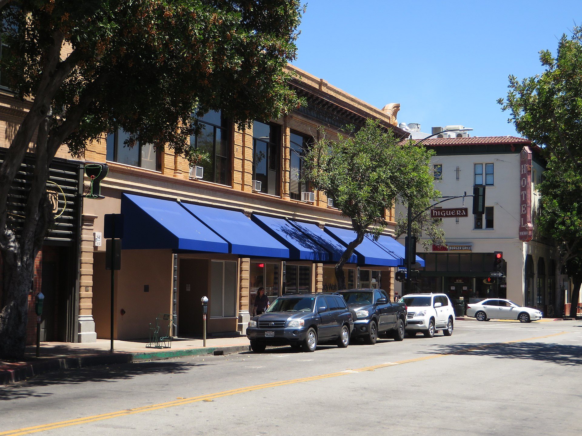 Alt text: Street view of a two-story building with blue awnings, parked cars, and a clear blue sky.