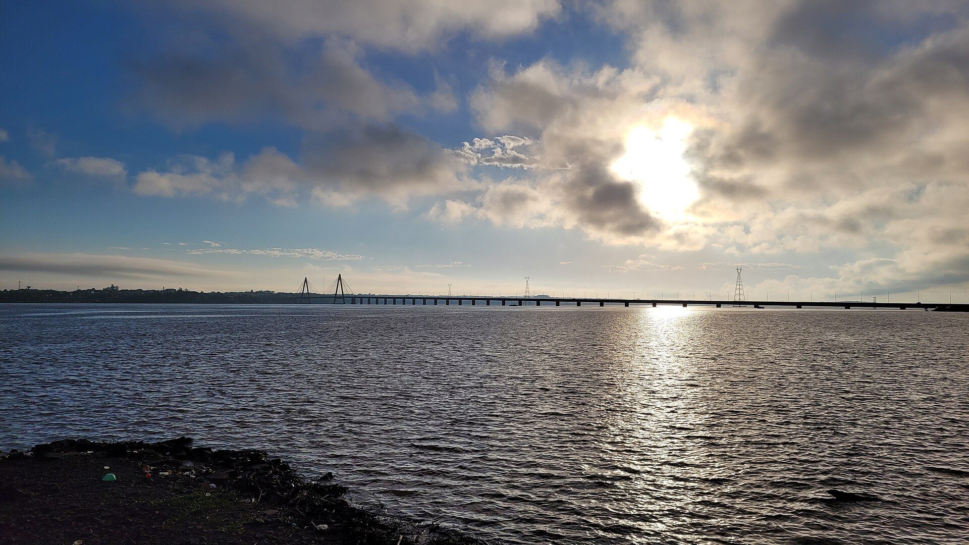 Alt text: "Panoramic waterfront view with a long bridge and partly cloudy sky.