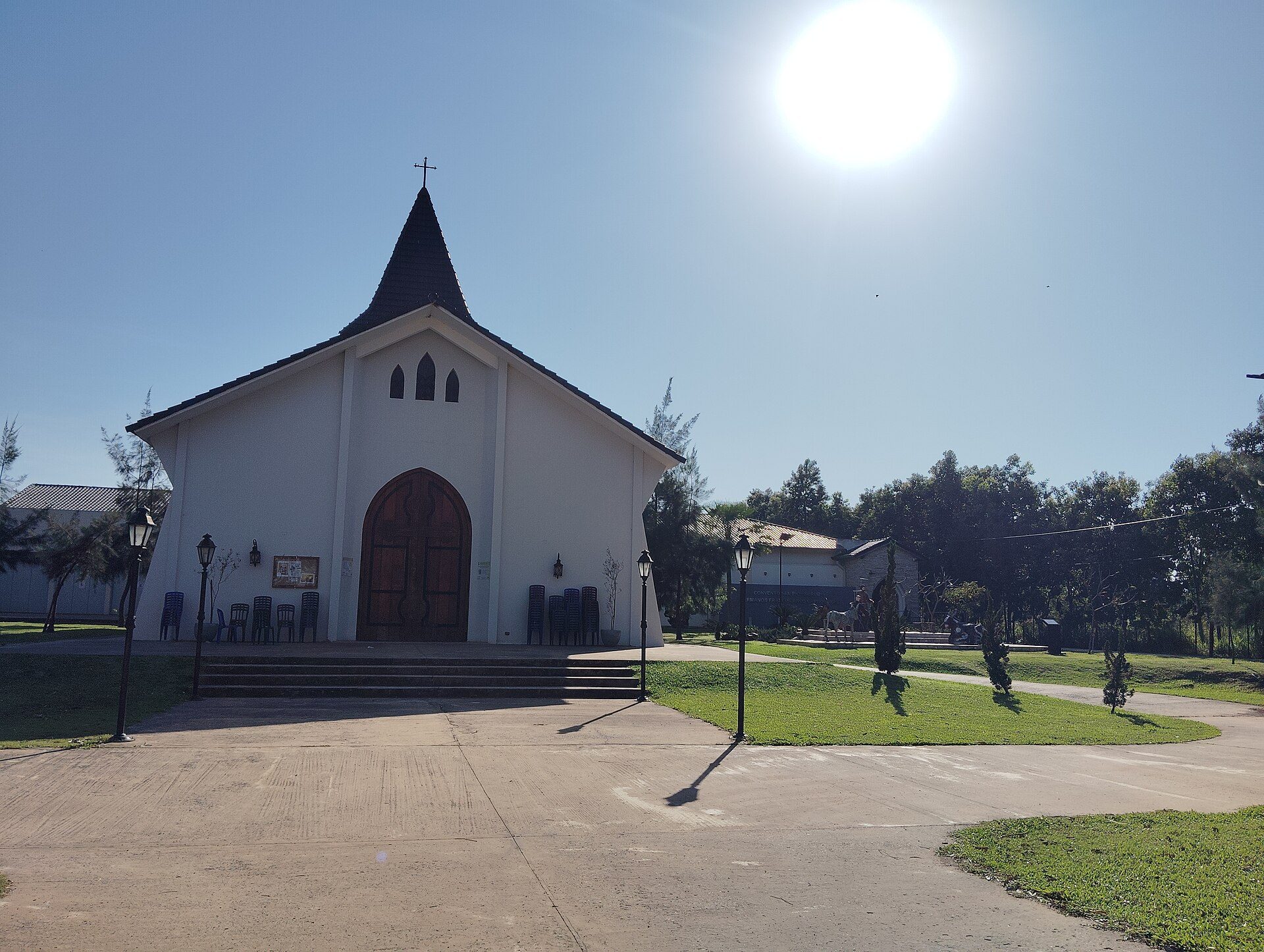 A small white church with a pointed roof and cross, surrounded by greenery and a clear blue sky.