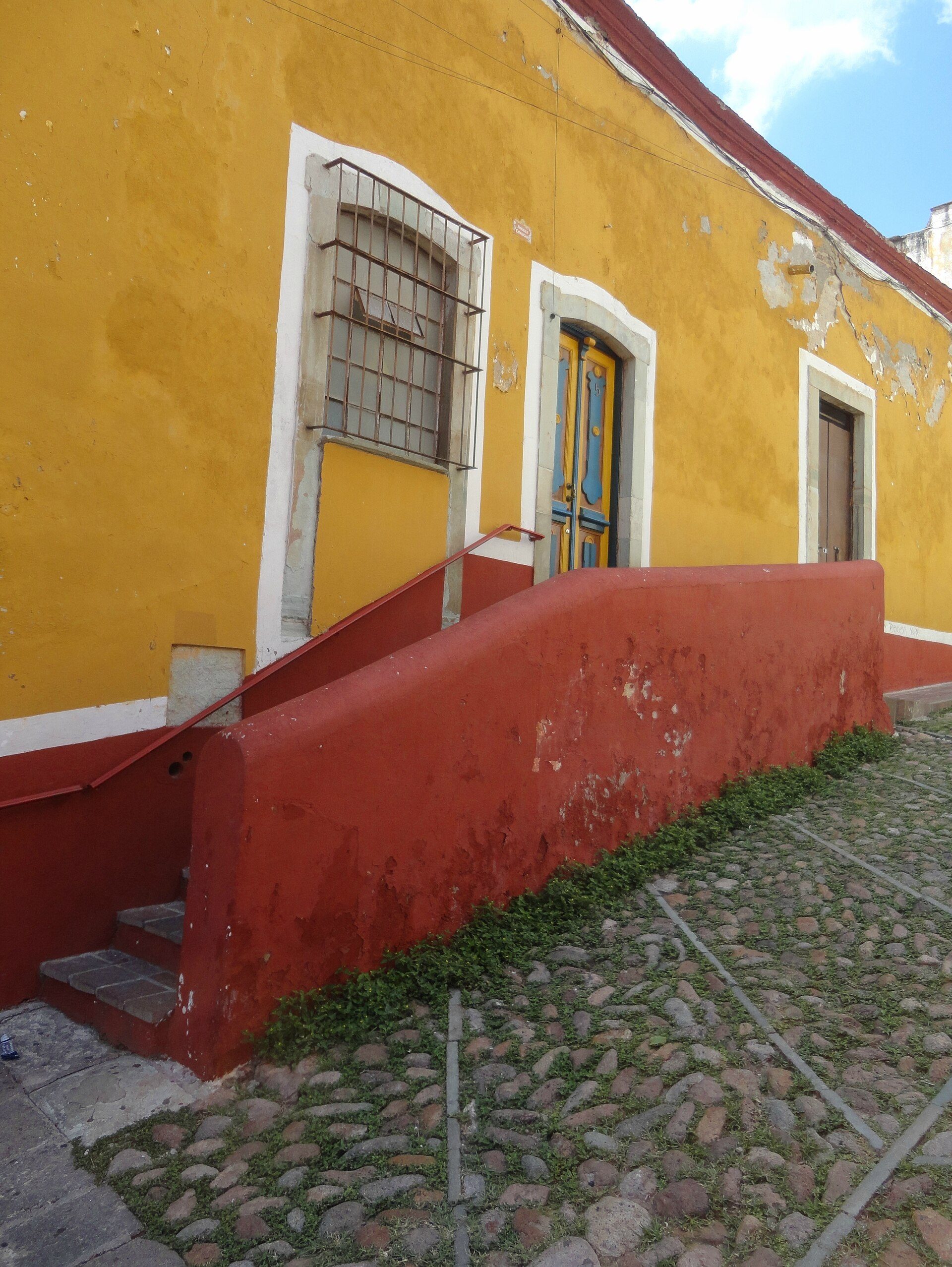Yellow building with red staircase, cobblestone path, barred window, and blue door.