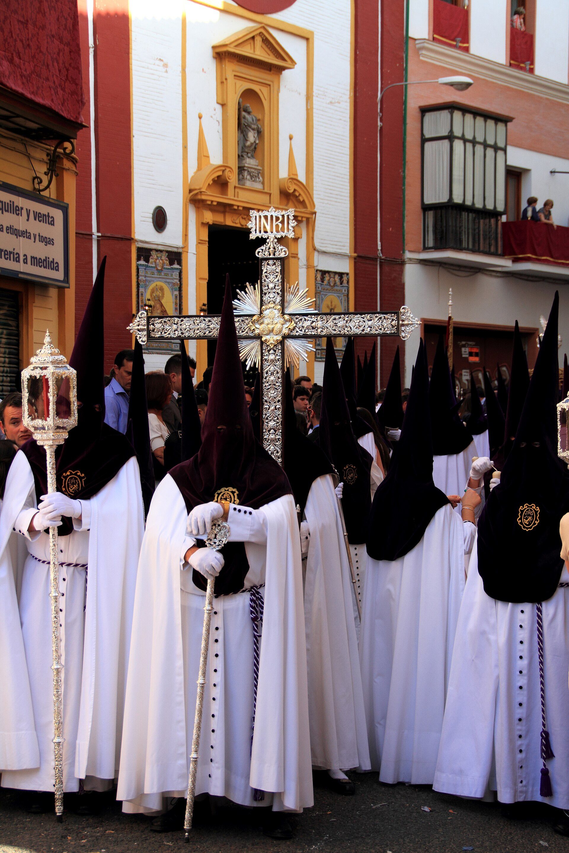 Religious procession with participants in white robes and black hoods, carrying a large cross, in front of a church.