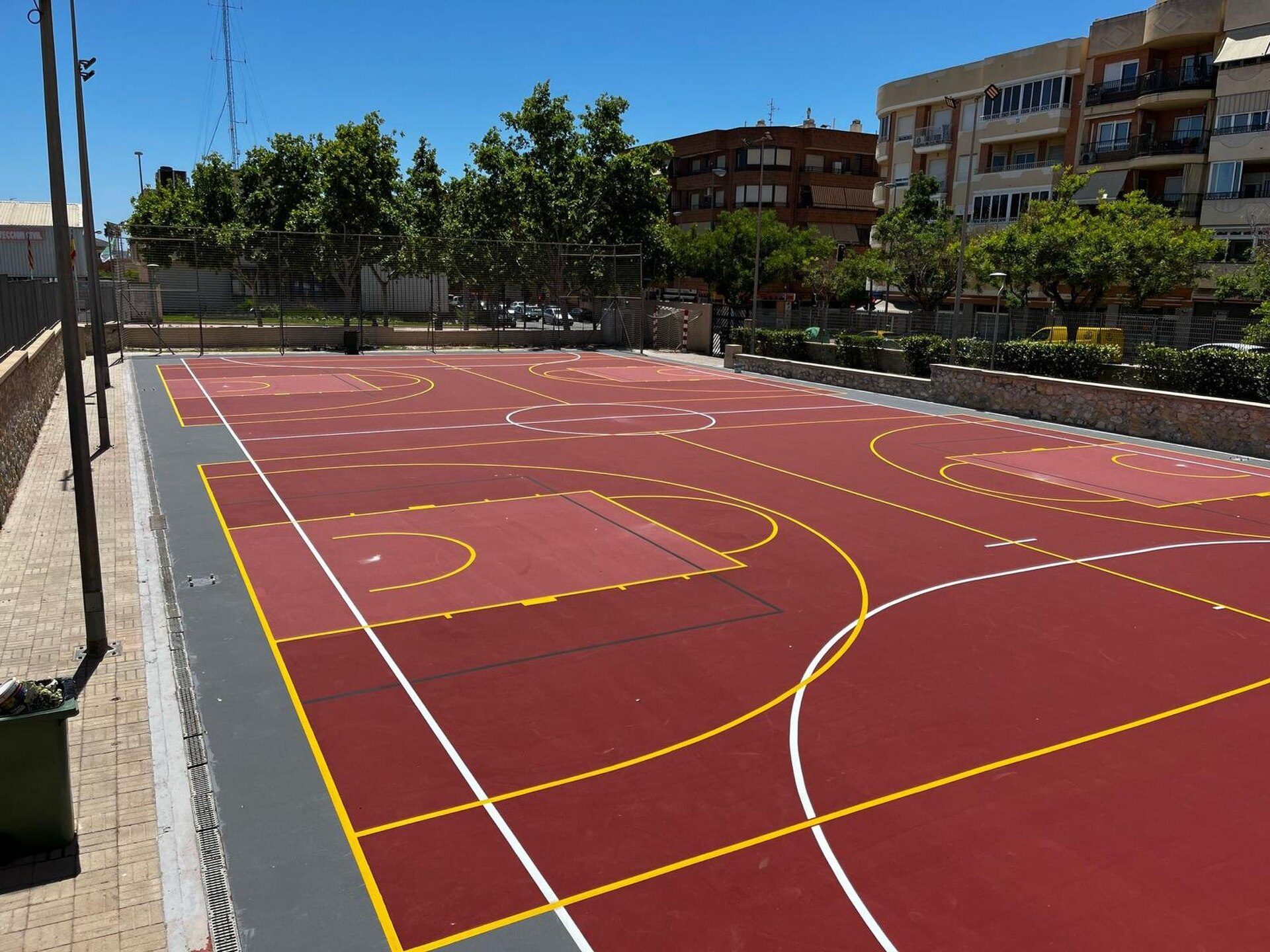 Outdoor basketball court with red surface, surrounded by trees and buildings.