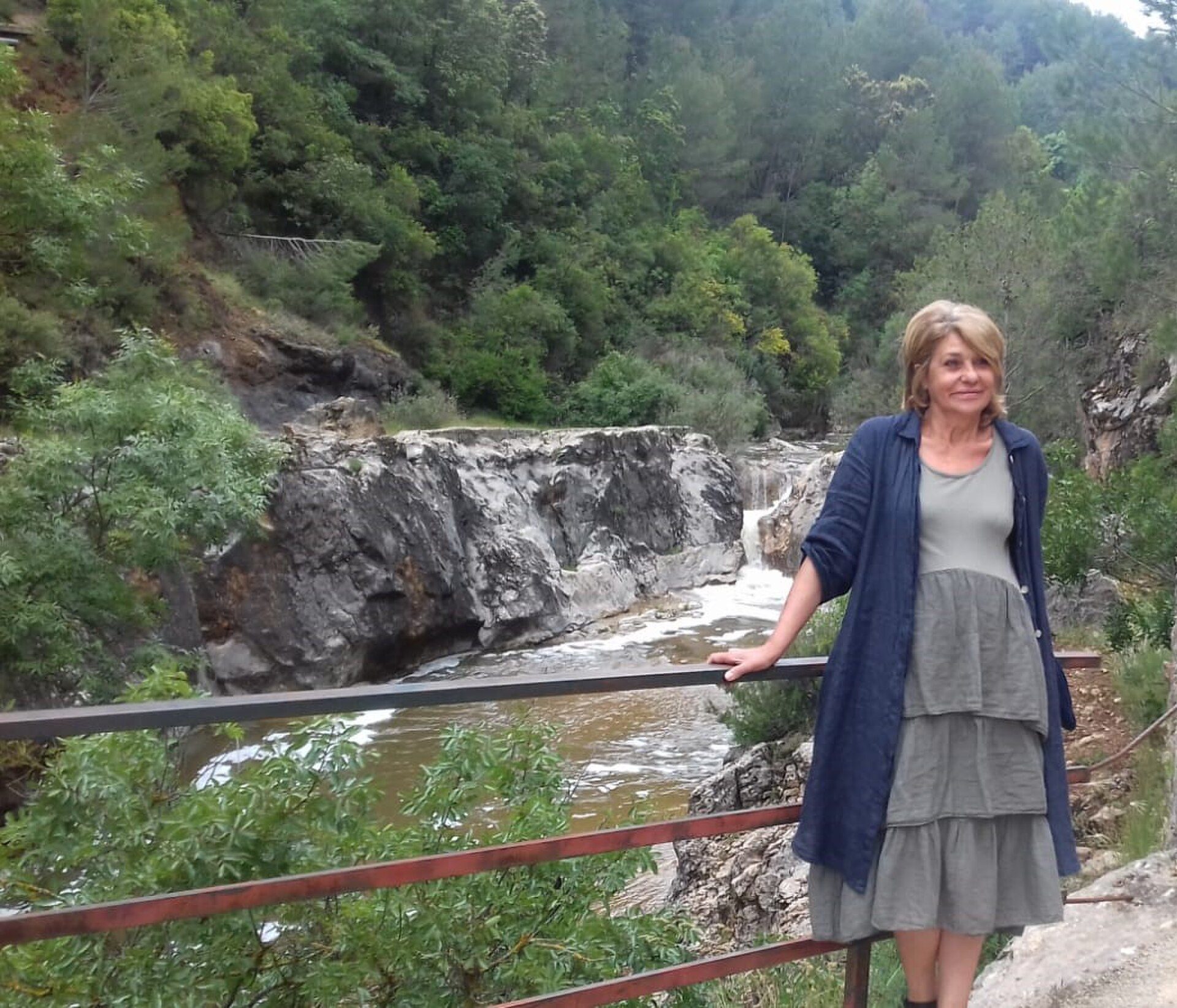 A woman stands on a balcony overlooking a scenic waterfall and lush greenery.