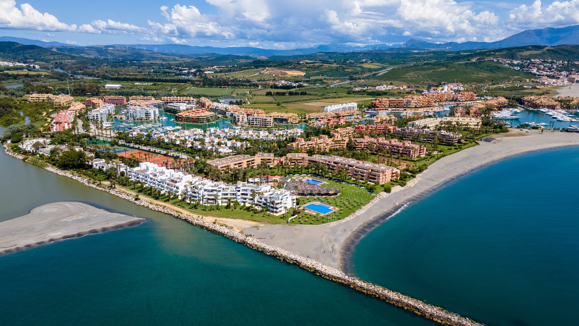 Aerial view of a coastal resort with multiple swimming pools, beach, and mountainous backdrop.