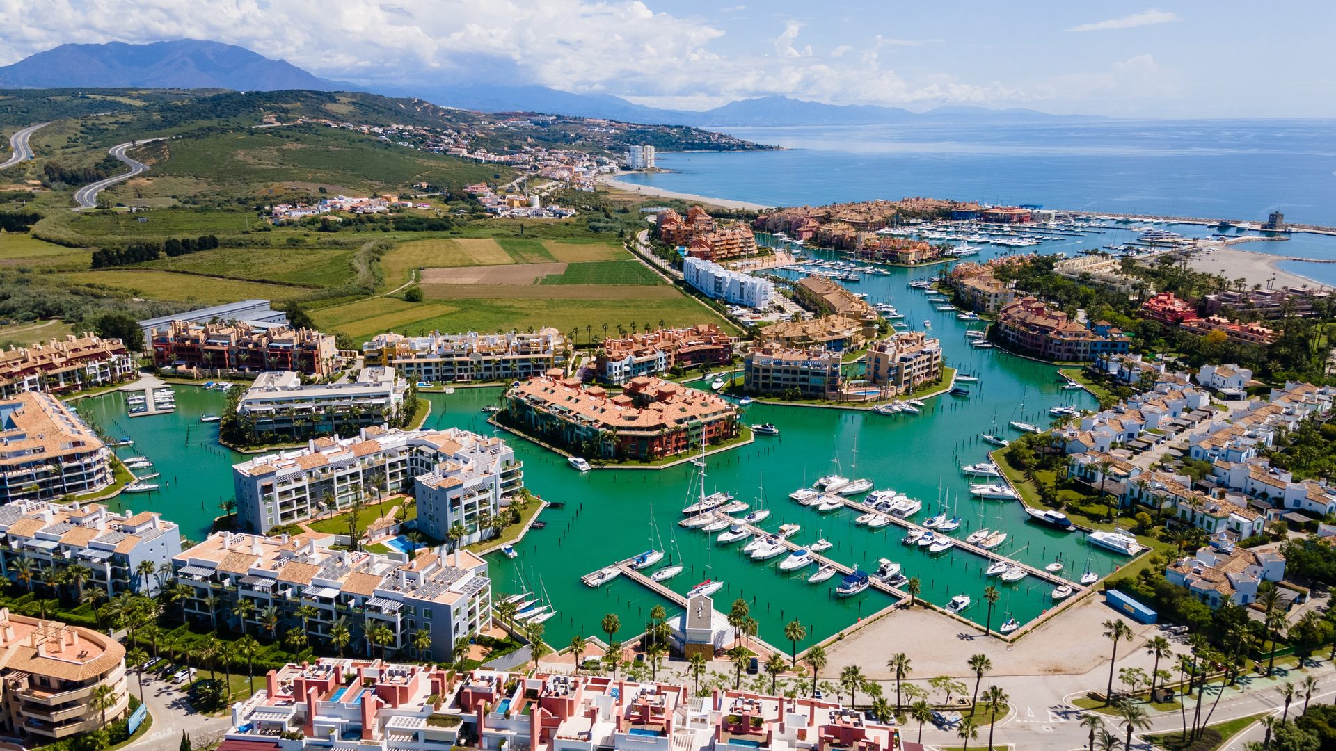 Aerial view of a coastal town with a marina, green fields, and mountains in the background.