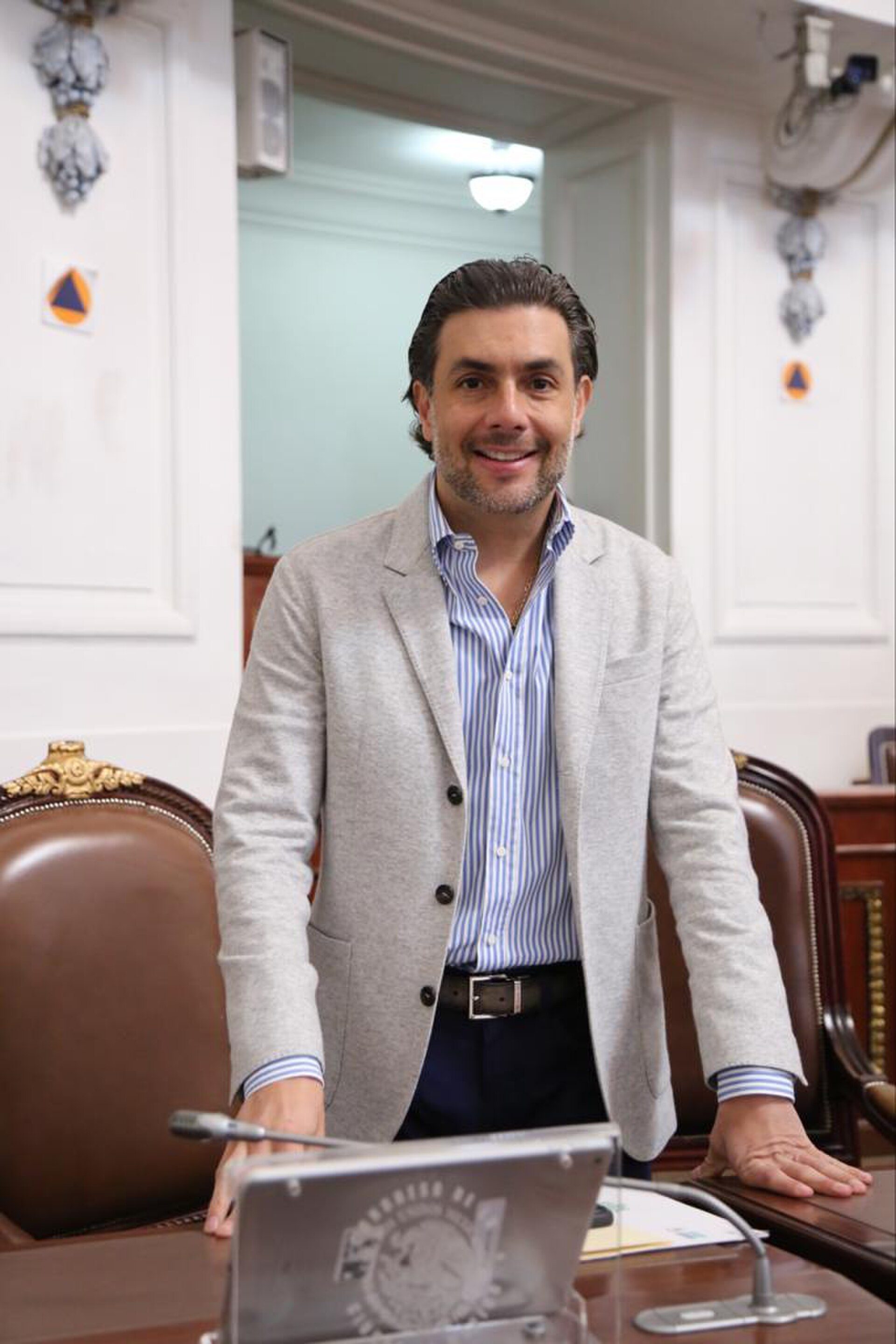 A man stands in an office with a laptop, featuring a formal setting with a chair and decorative elements.
