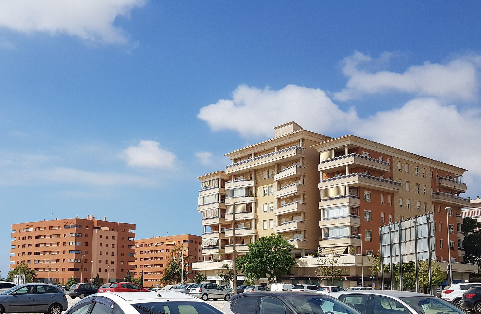 A modern apartment complex with balconies, offering city views, and surrounded by parked cars.