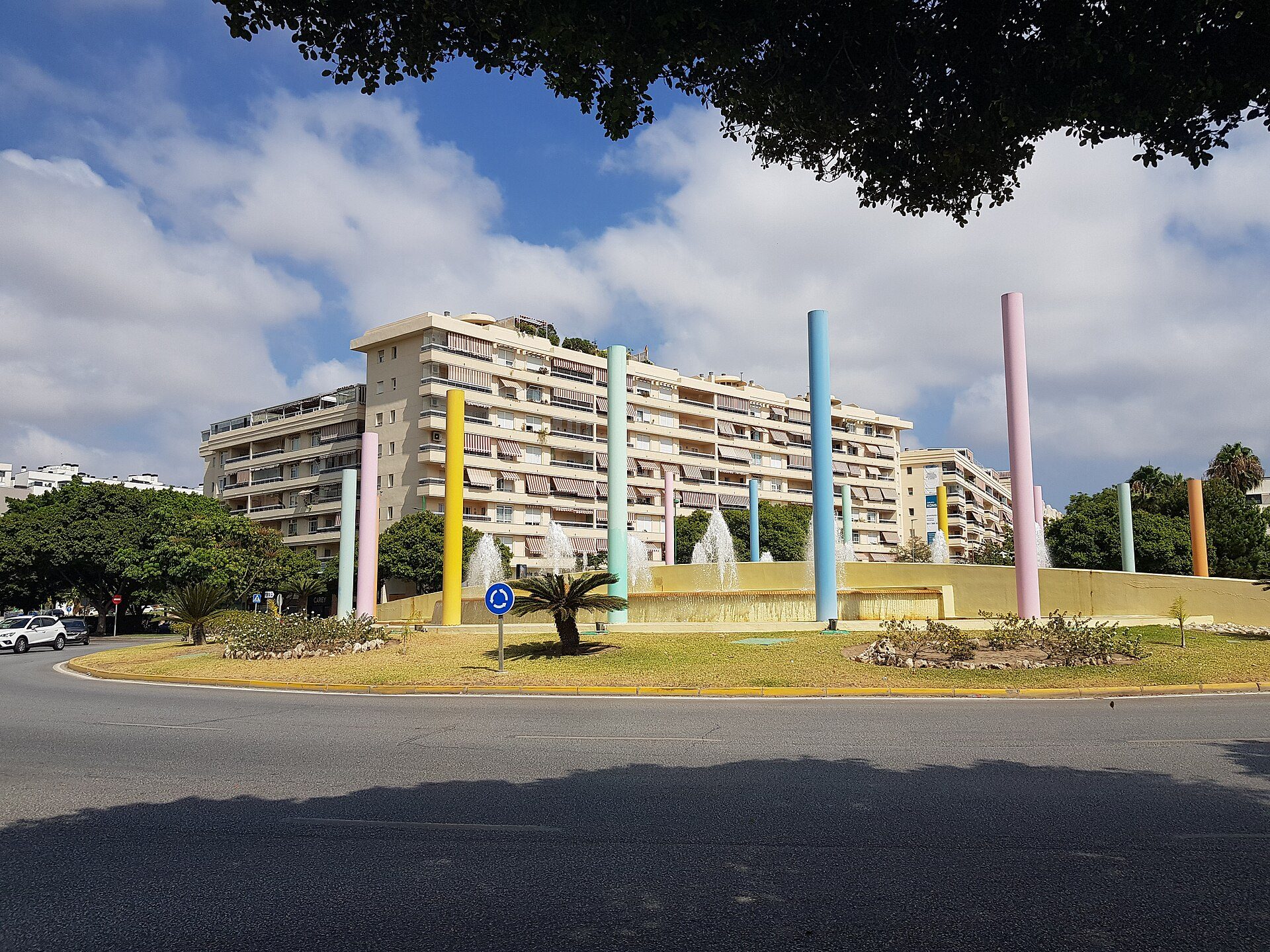 Alt text: Colorful fountain in front of modern apartment building with balconies and greenery.