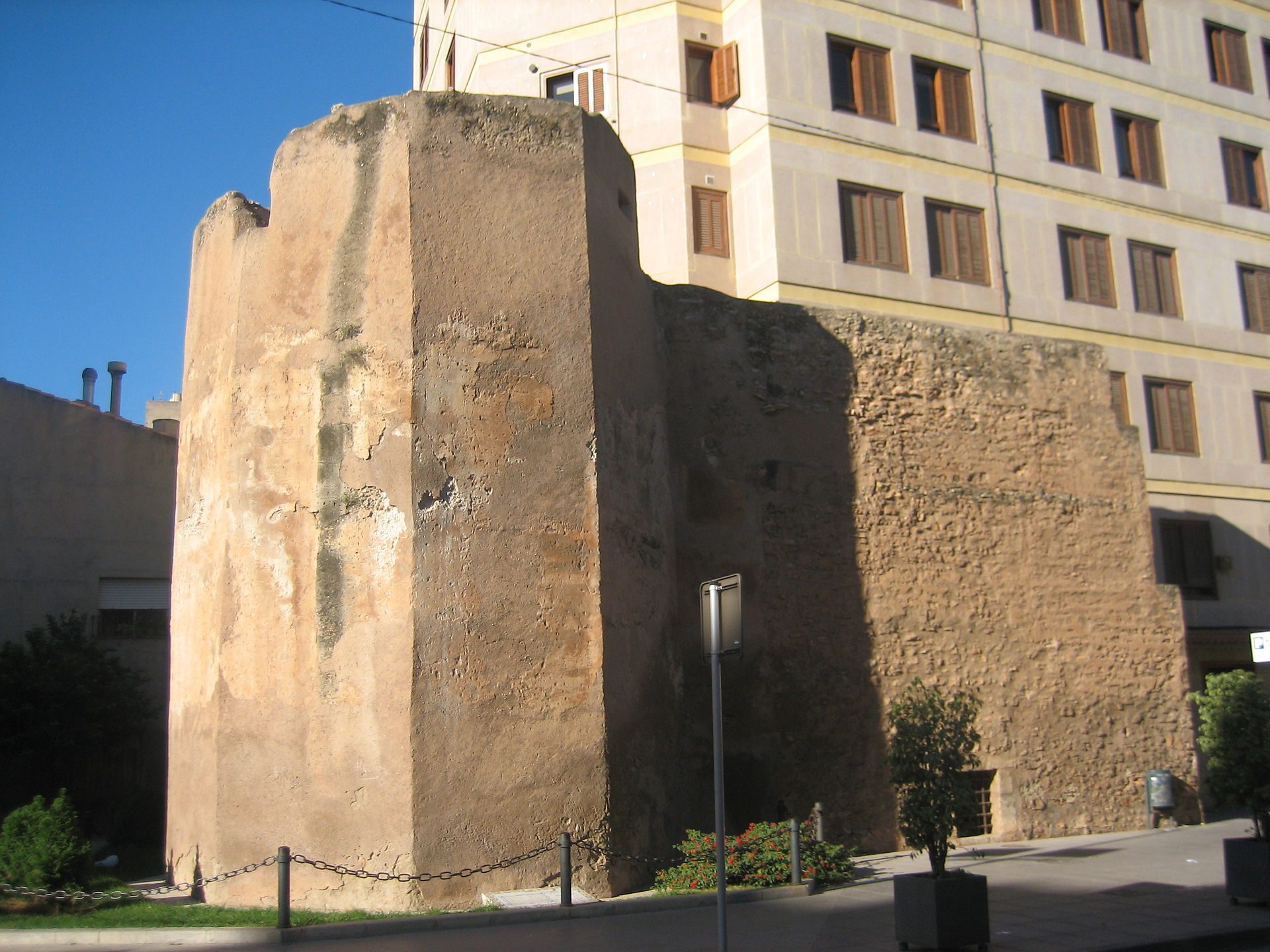 A historic stone tower with a modern building in the background, surrounded by greenery.