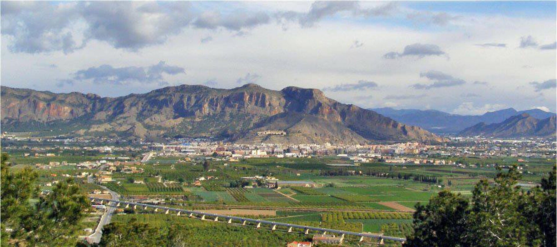 Alt text: Panoramic view of a mountainous landscape with a city and green fields.