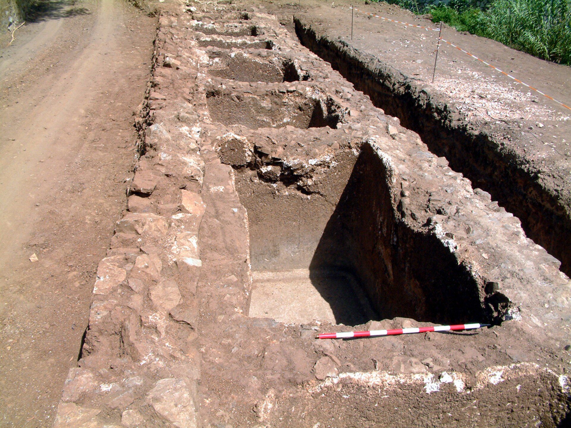 An archaeological site with multiple rectangular pits, surrounded by a dirt path and vegetation.