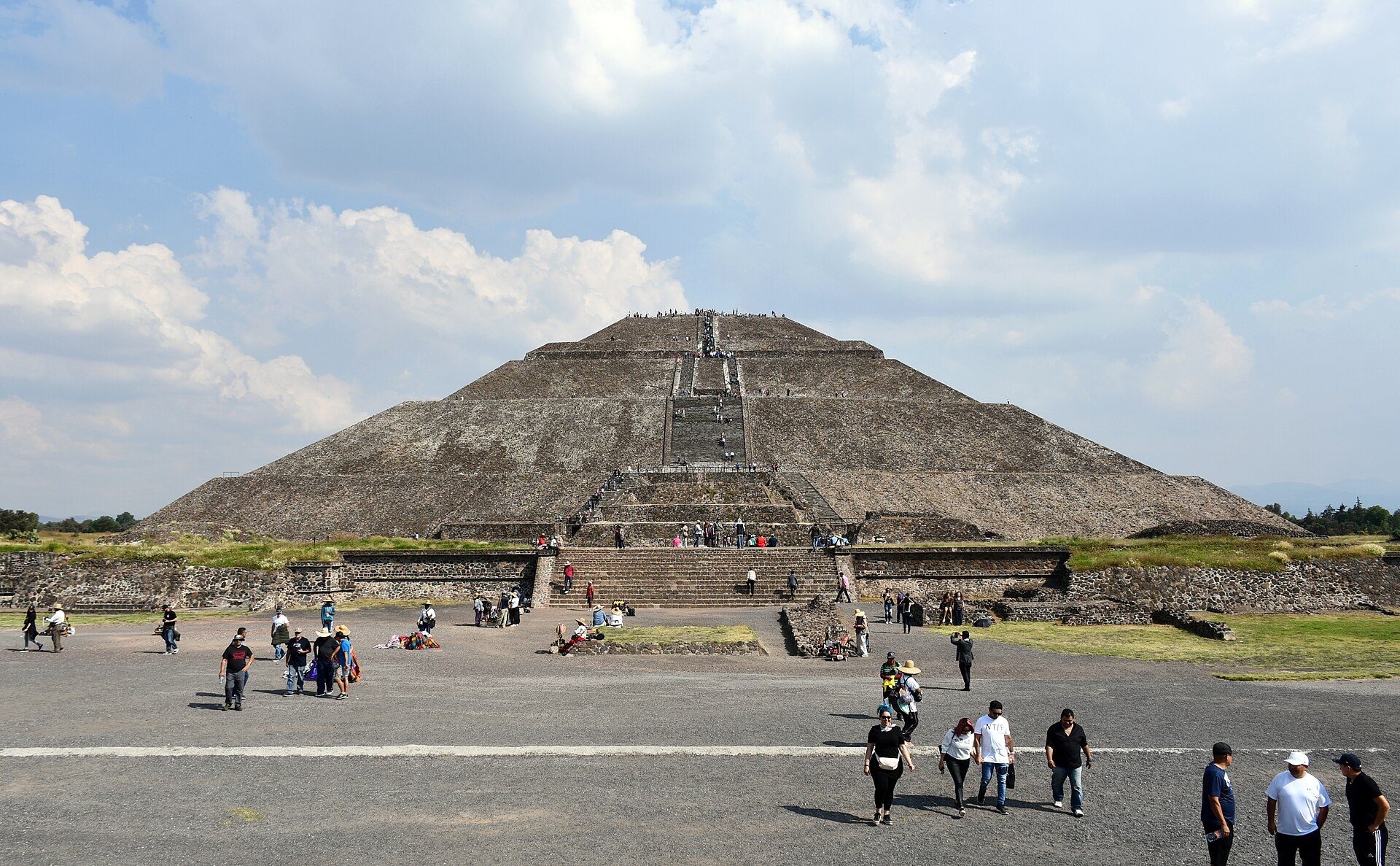 Alt text: Historic pyramid with steps, surrounded by visitors under a partly cloudy sky.