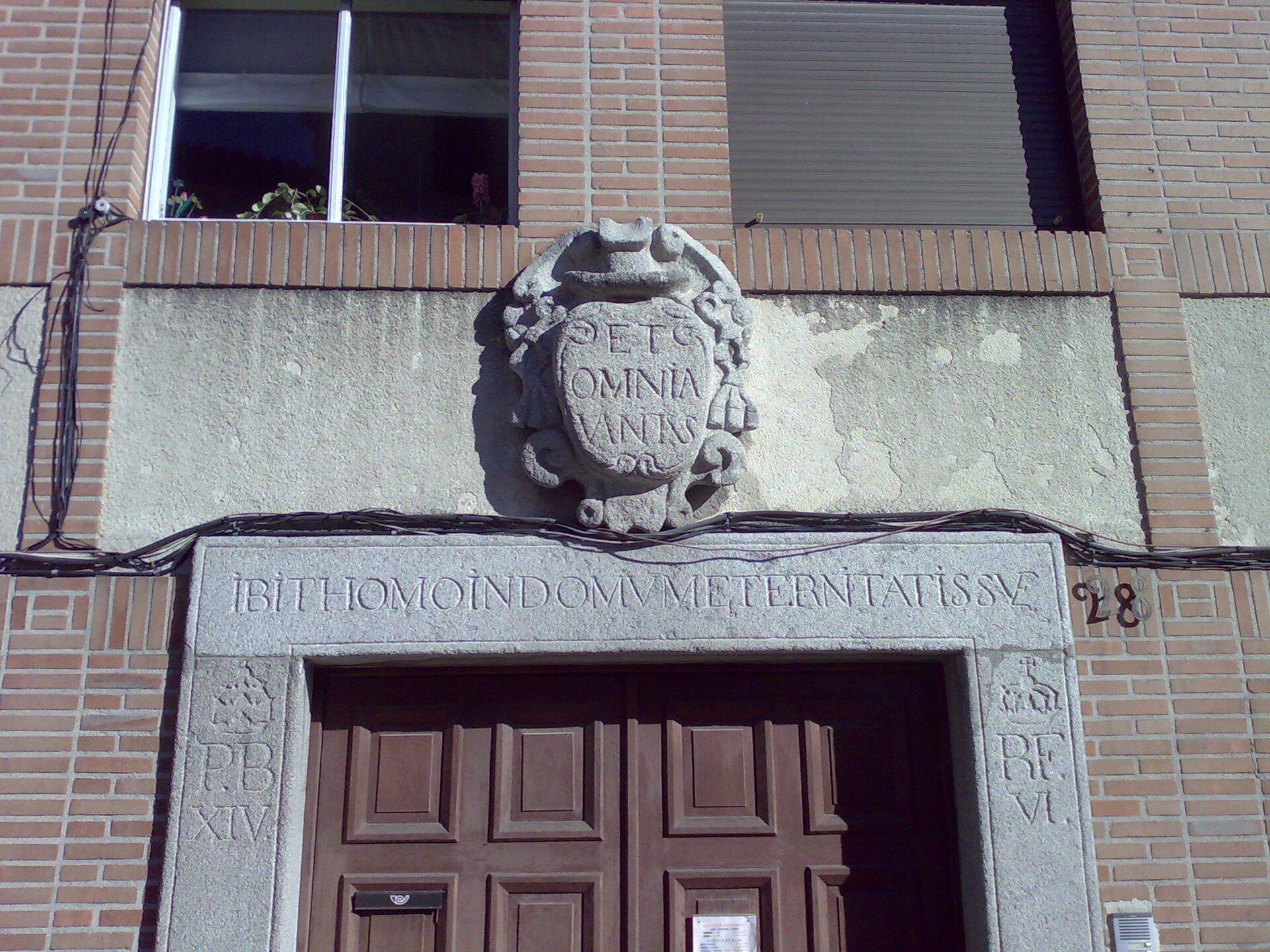 Alt text: Stone facade with Latin inscription, wooden double doors, brick building, window with plants.