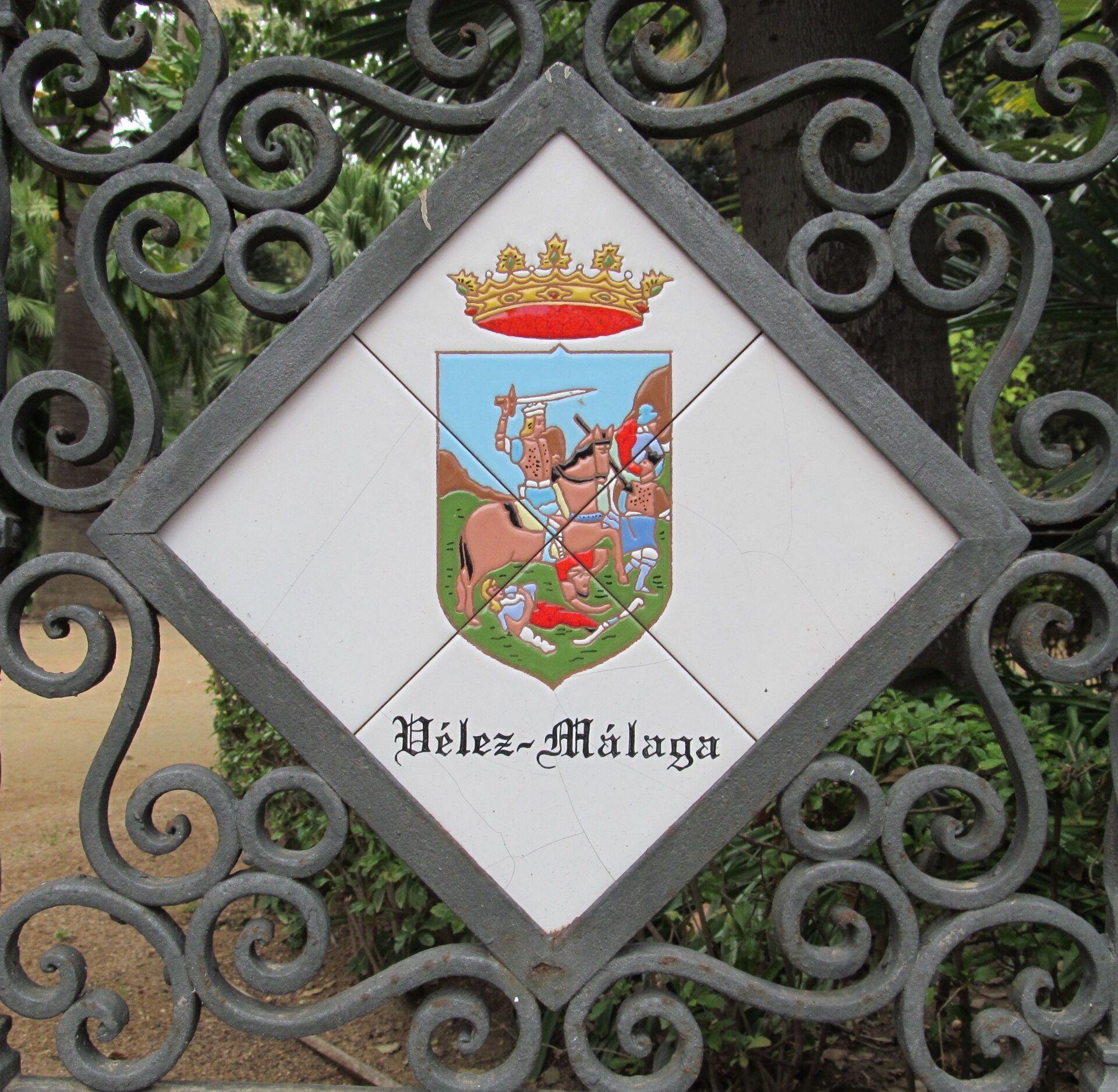 Diamond-shaped sign with coat of arms and "Hélez - Málaga" text, ornate metal gate, greenery background.