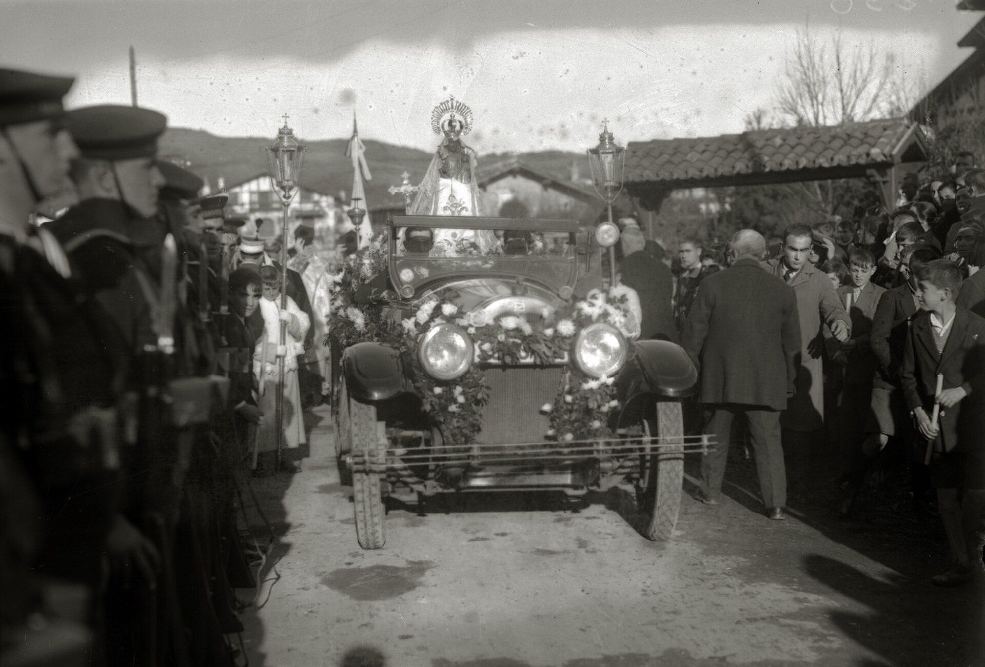 Alt text: Vintage black and white photo of a procession with a decorated car and people in formal attire.