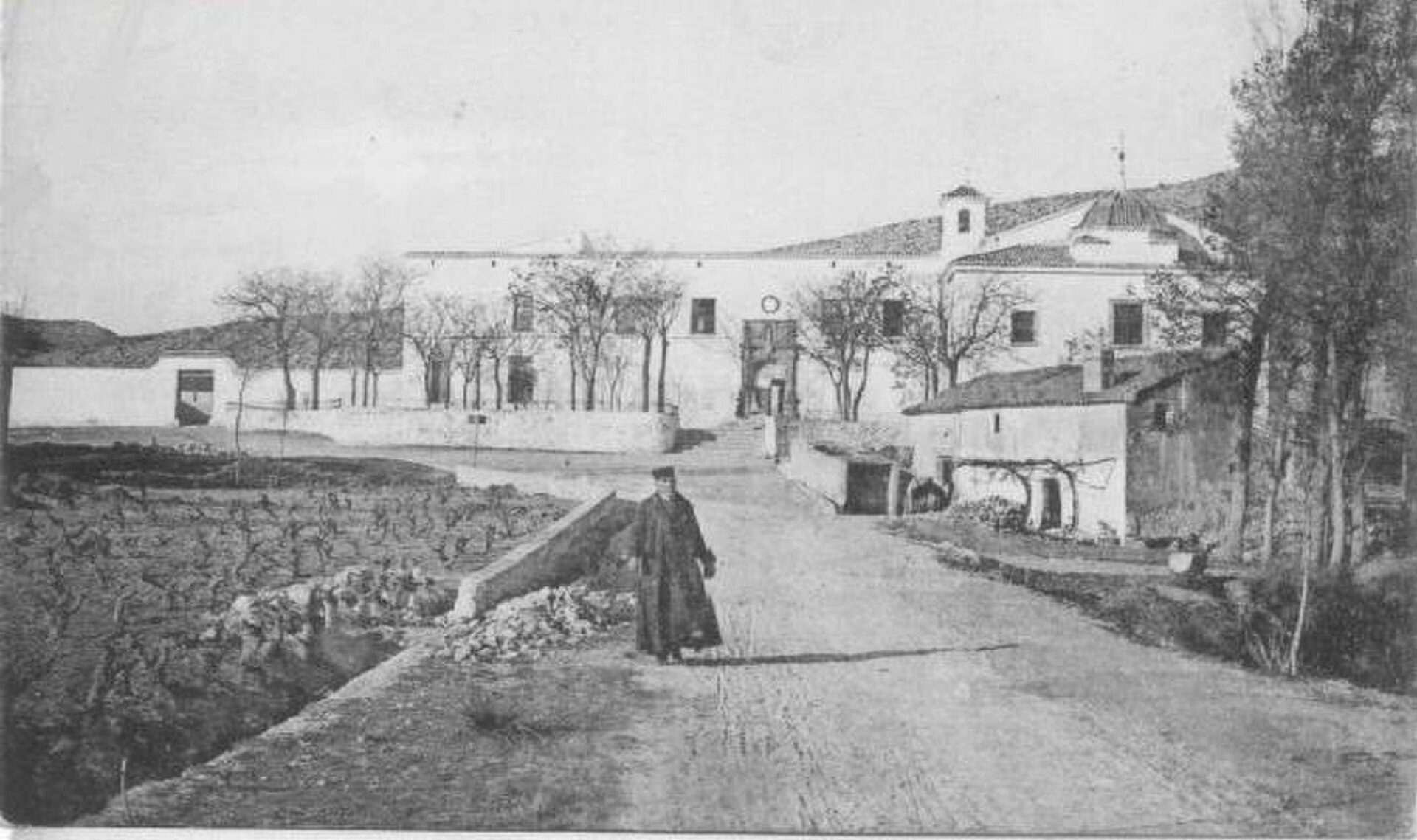 A black and white photo of a rural property with a courtyard, trees, and a person in traditional attire.