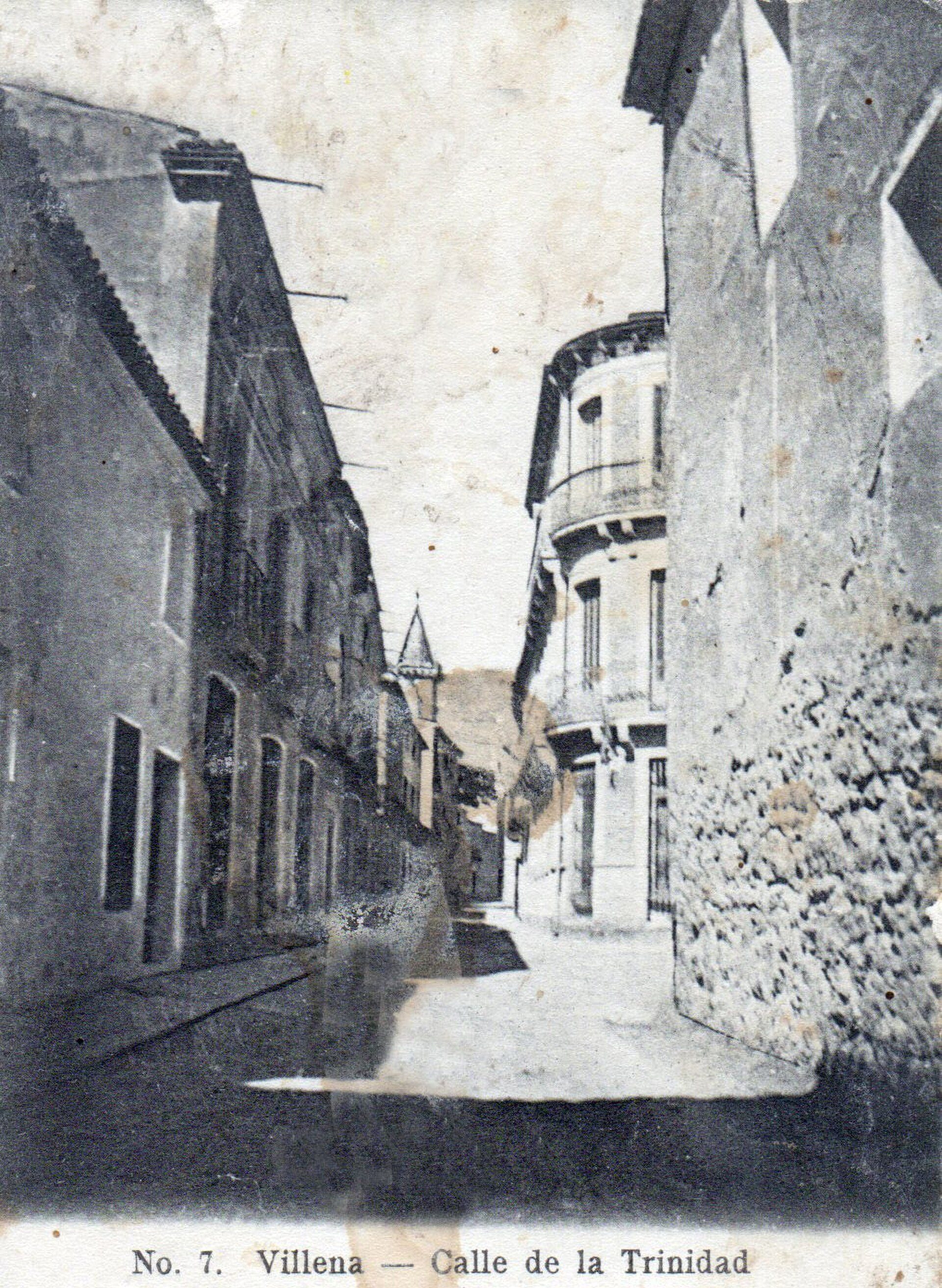 Black and white photo of a narrow street in Villena, Spain, with old buildings and a distant church.