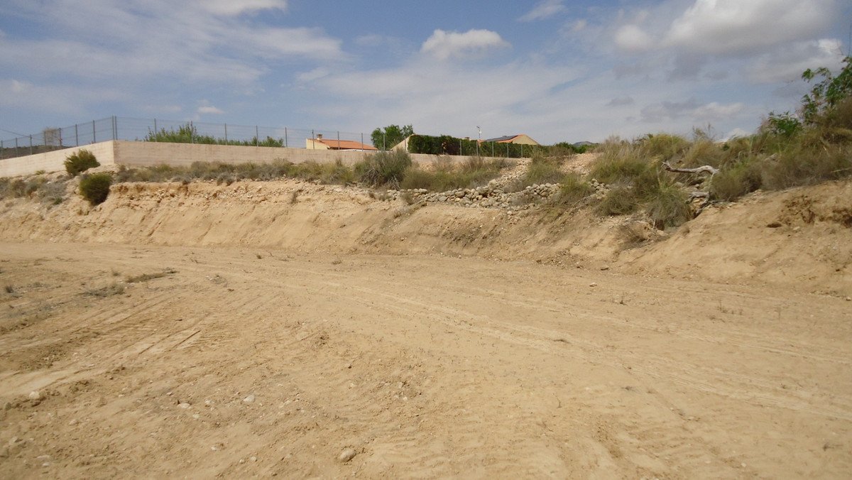 A wide dirt track leads through a dry, arid landscape towards houses visible in the distance, under a cloudy sky.