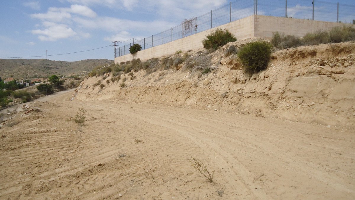 A sandy dirt road curves up a dry, barren hillside with scrubby bushes and a distant fence line.