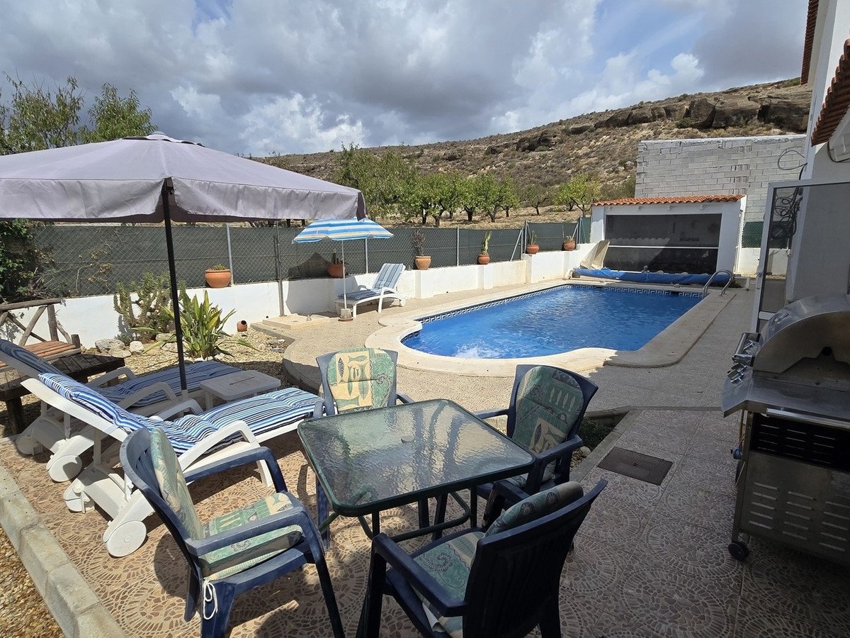 Outdoor patio with a swimming pool, lounge chairs, dining table, and a grill, set against a backdrop of hills.