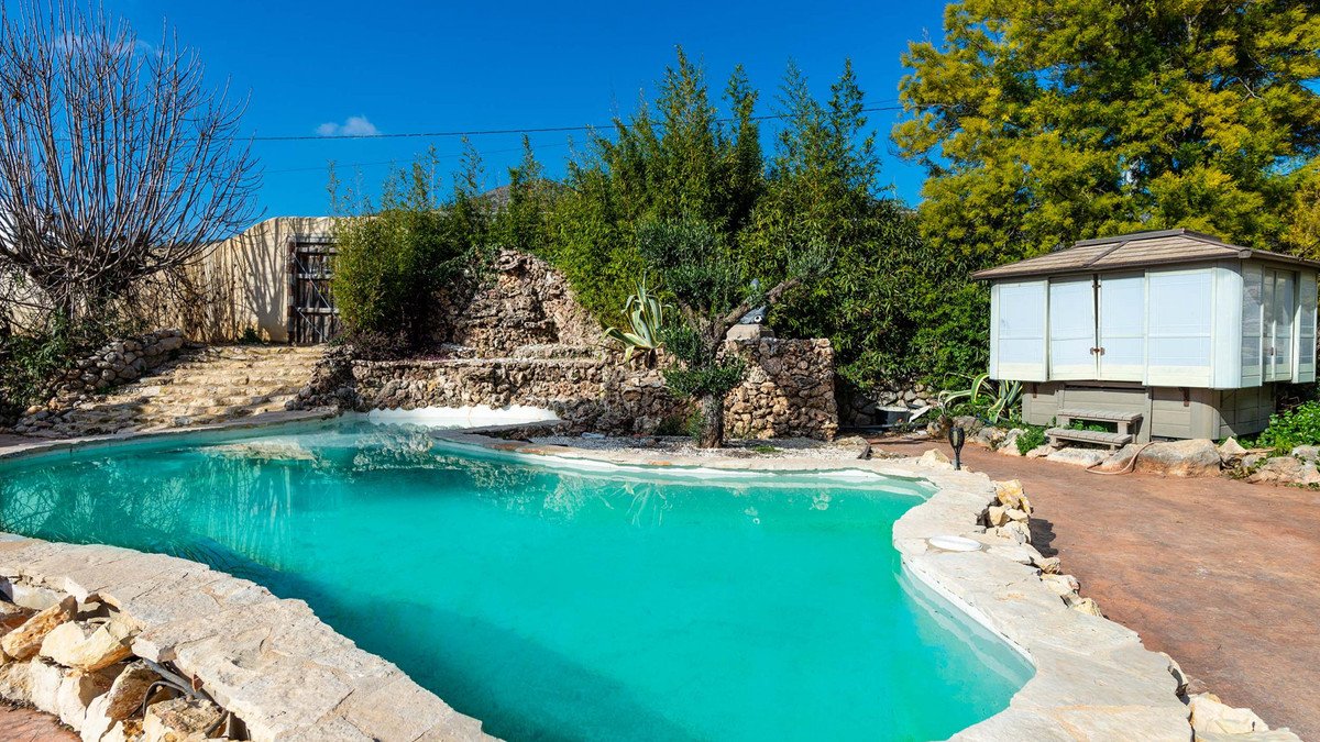 An inviting turquoise swimming pool with a natural stone waterfall feature and a modern gazebo in a lush garden setting.