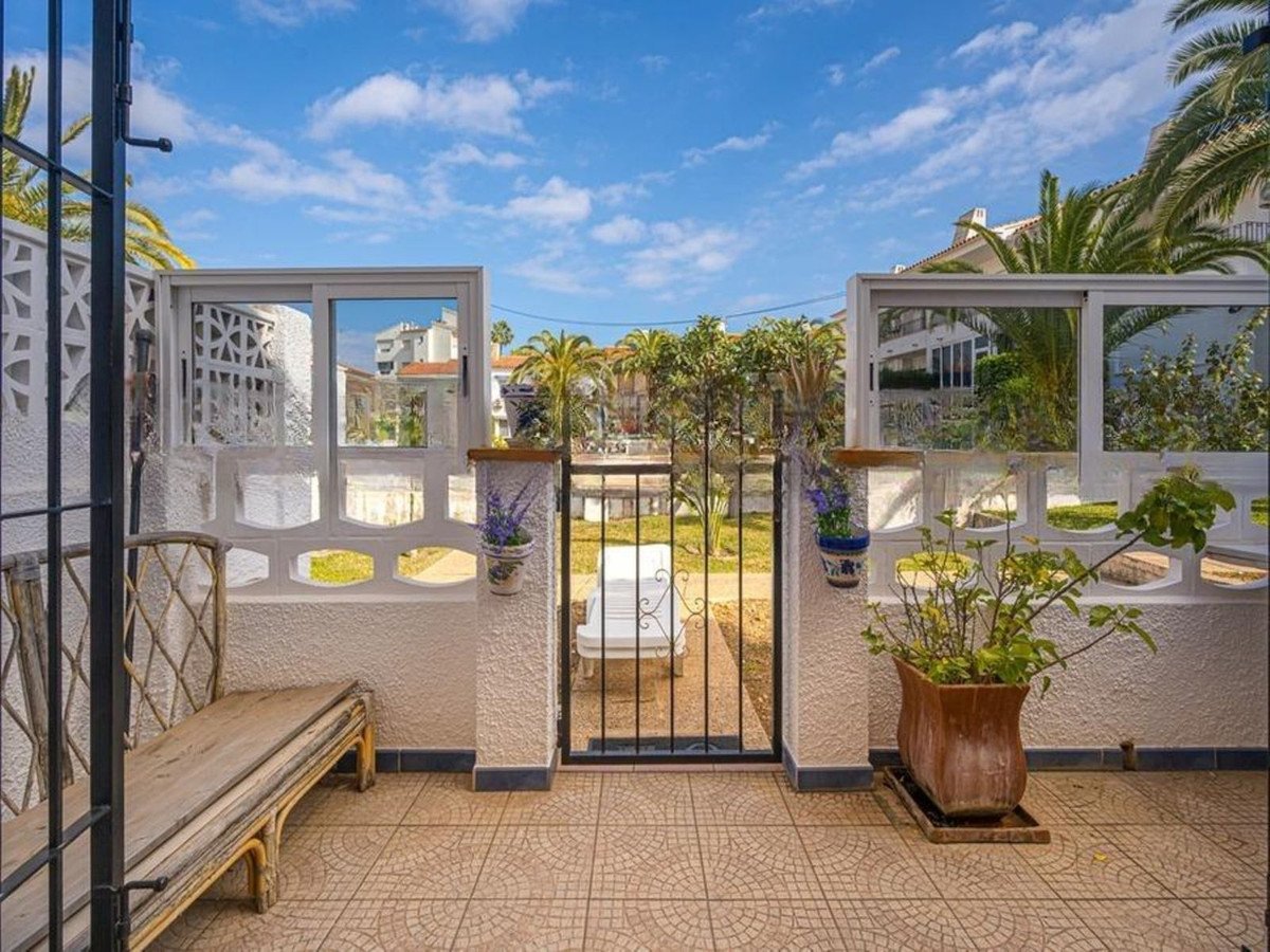 A patio with a wooden bench, a gate leading to a garden, and potted plants under a blue sky.