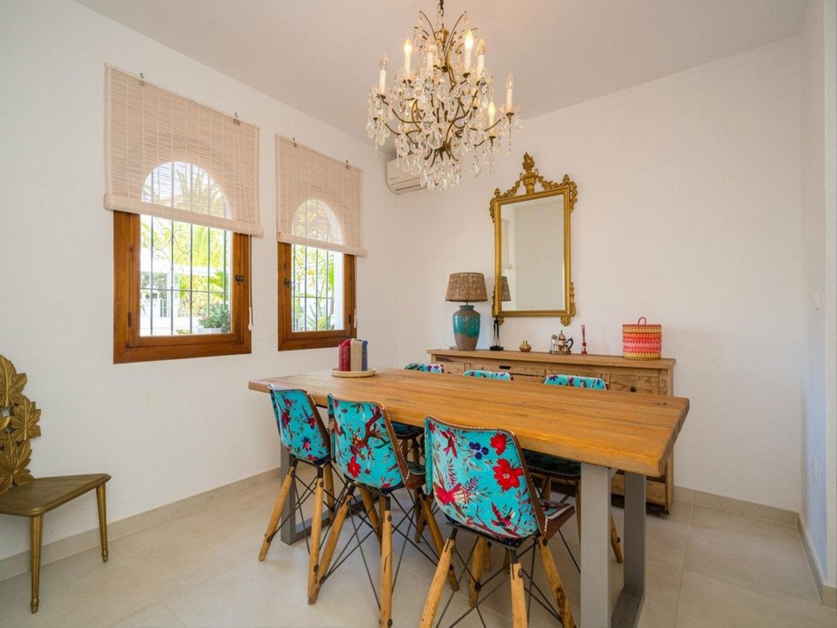 Dining room with a wooden table, colorful floral chairs, a crystal chandelier, and a decorative mirror above a wooden cabinet.