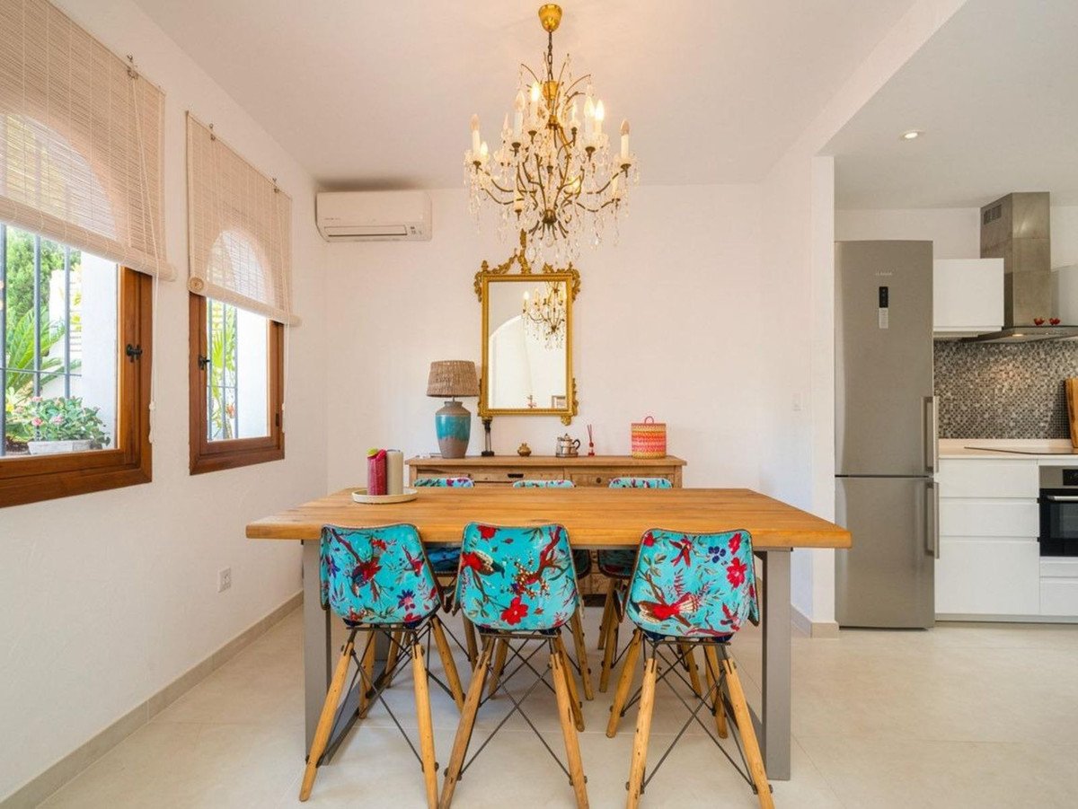 A dining area with a wooden table, colorful chairs, and a crystal chandelier, adjacent to a kitchenette with a refrigerator.
