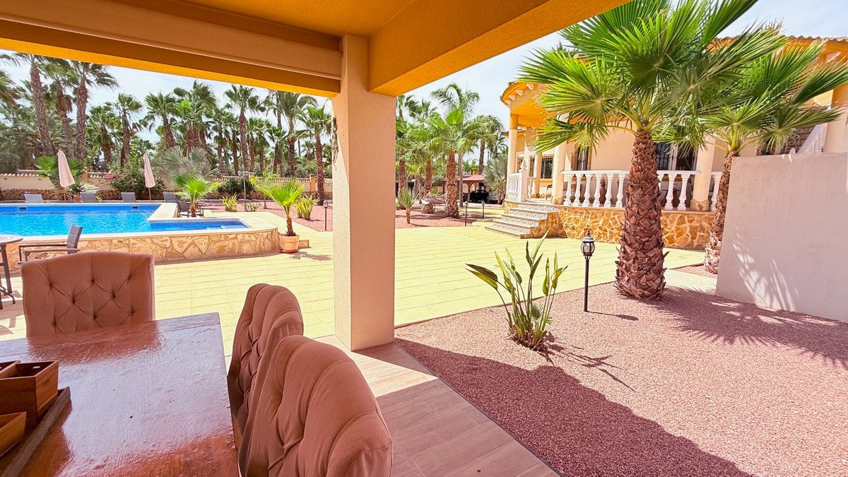 Covered patio with dining table and chairs overlooking a pool and lush garden with palm trees.