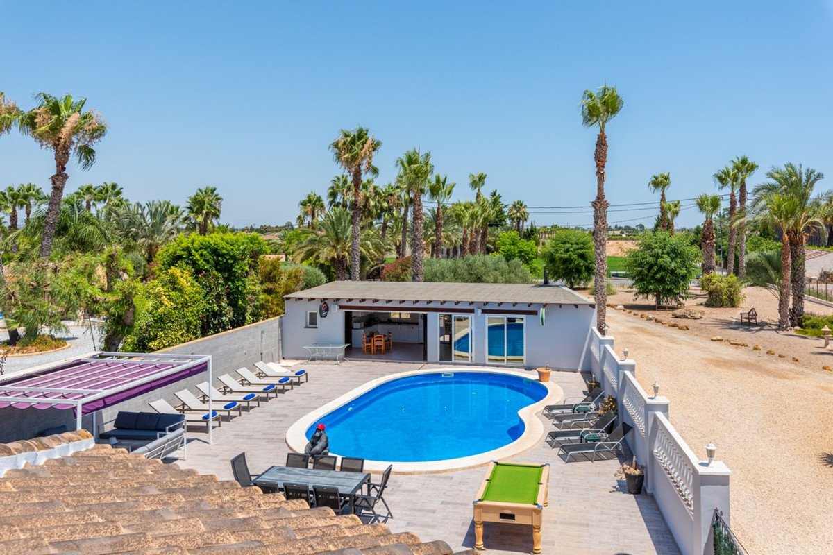 A spacious outdoor area with a swimming pool, lounge chairs, dining table, and pool table, surrounded by palm trees.