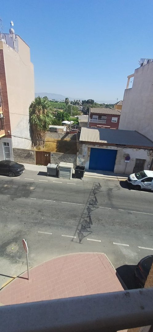 Balcony view of a street with buildings, palm trees, and distant mountains under a clear blue sky.