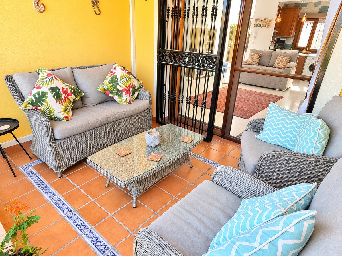 Covered terrace with comfortable wicker seating, tropical and chevron patterned cushions, and a glass coffee table, opening to an interior living area.
