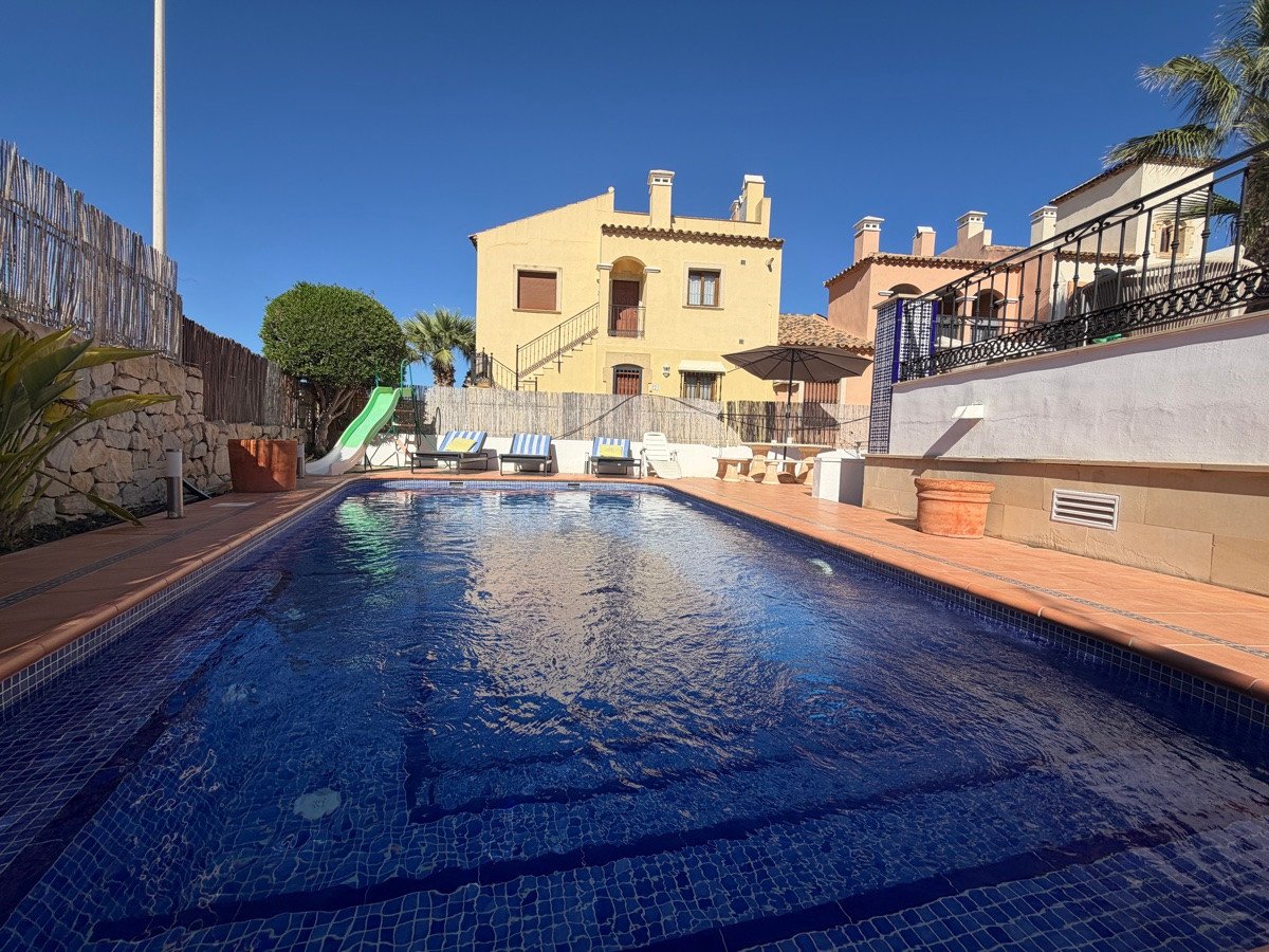 A sparkling blue swimming pool with a green slide and sun loungers, surrounded by houses and a patio area.