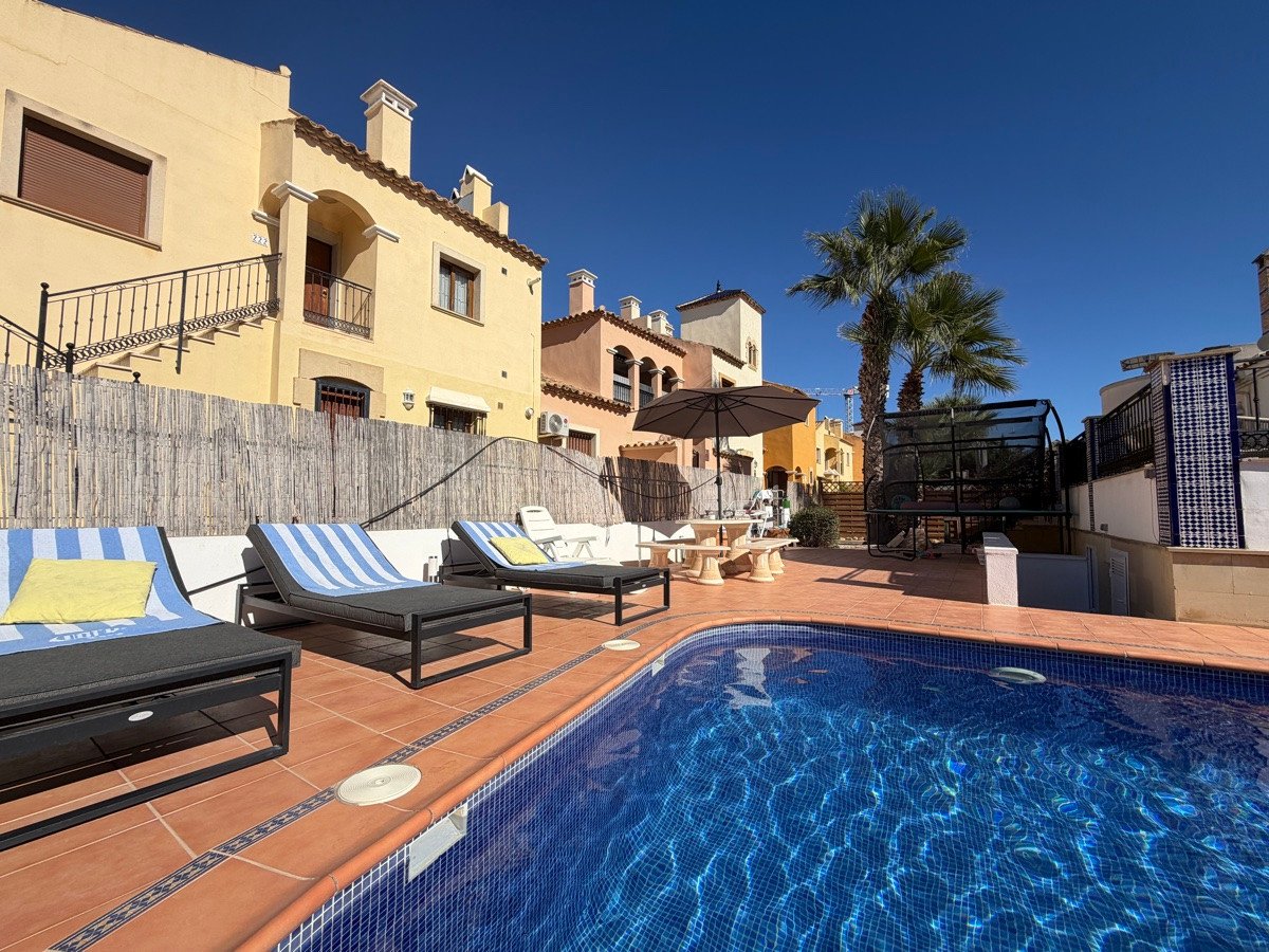 Exterior view of a property featuring a private swimming pool, lounge chairs, a table with an umbrella, and palm trees under a clear blue sky.