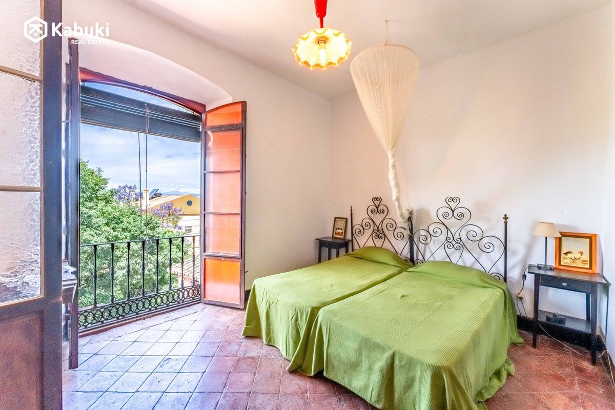 Bedroom with two beds, a balcony with a view of trees and houses, and antique-style metal headboards.
