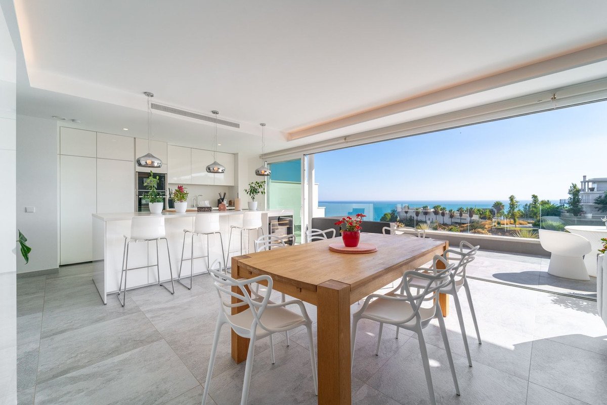 Modern kitchen and dining area with bar stools, leading to a balcony with a wooden table and a stunning sea view.