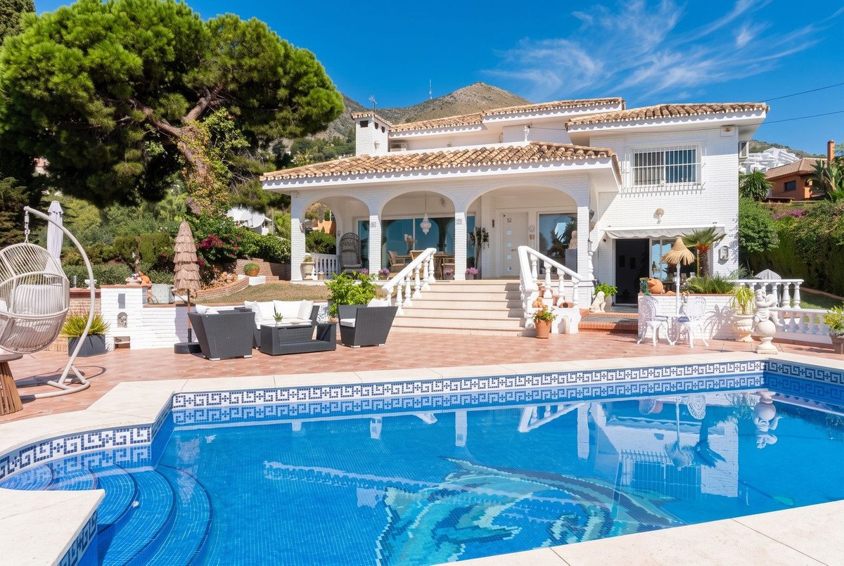 A beautiful white villa with a swimming pool, patio furniture, and lush greenery, offering stunning views.