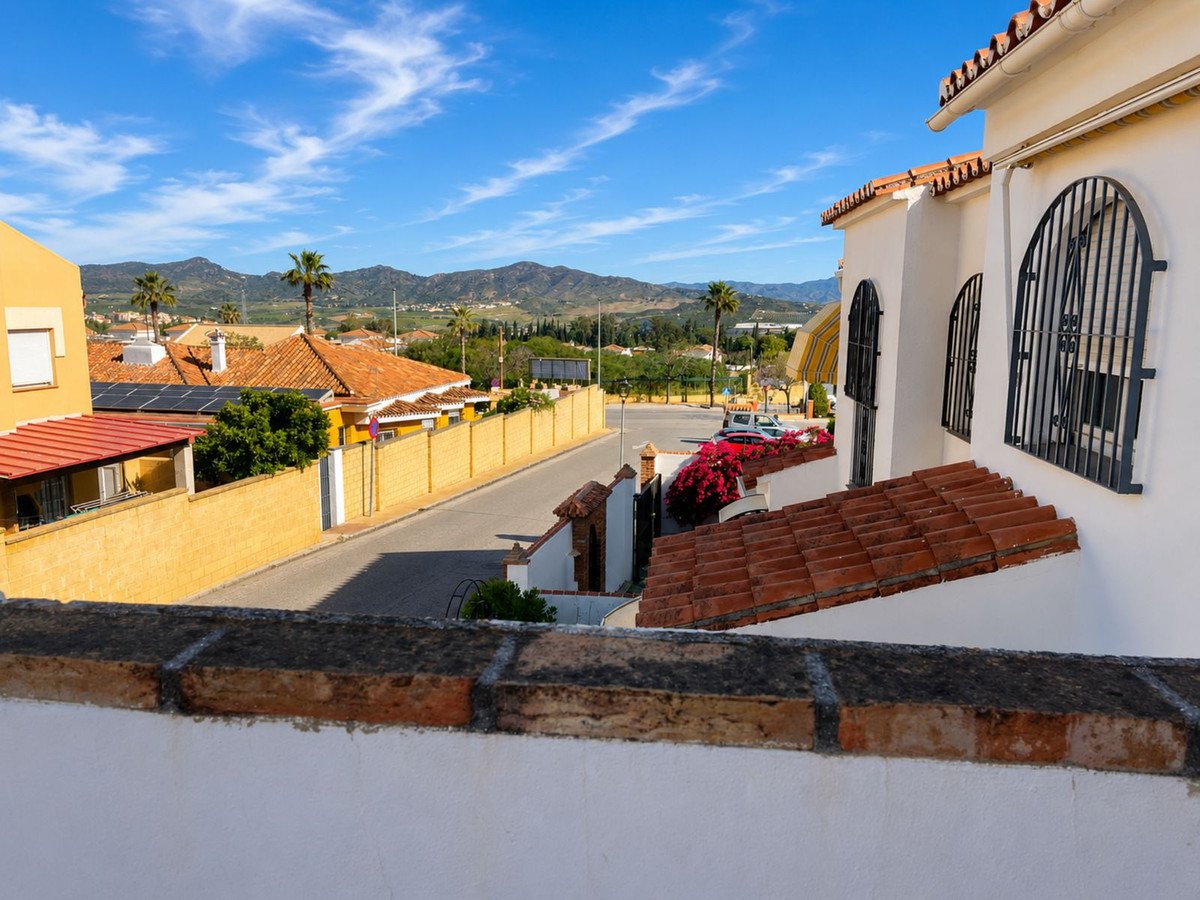 Balcony view of a white villa with terracotta roof, overlooking a street and distant mountains under a blue sky.