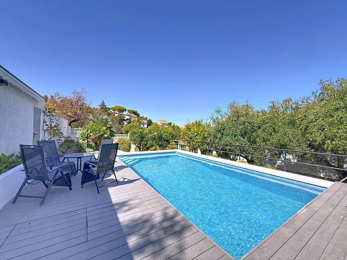 Dæk ved poolen med solstole, klar blå pool og frodige grønne træer under solrig himmel.