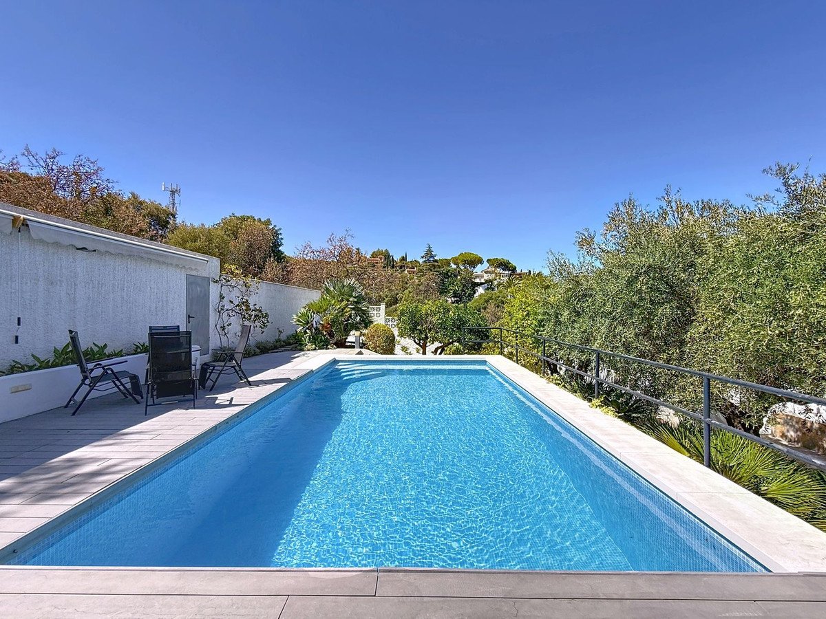 Rektangulær pool med klar blåt vand omgivet af patio og frodig natur under solrig himmel.