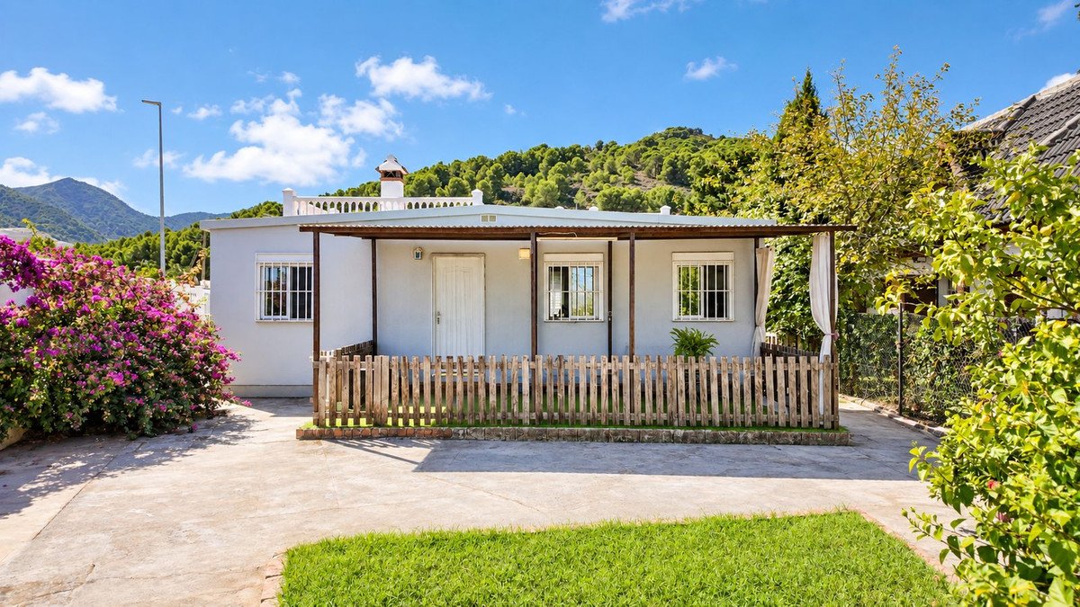 Small cottage with a covered porch, wooden fence, and lush greenery against a backdrop of lush green mountains.