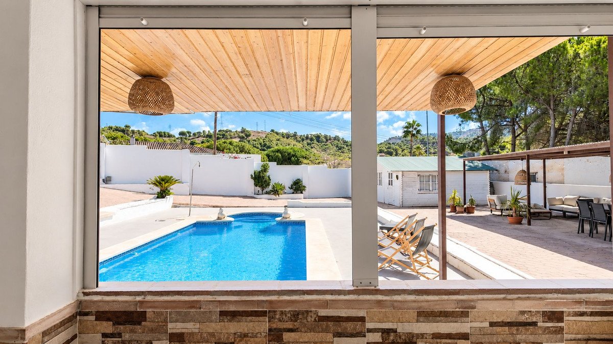 View of a bright blue swimming pool and a patio with outdoor seating and dining areas, set against a backdrop of green hills.
