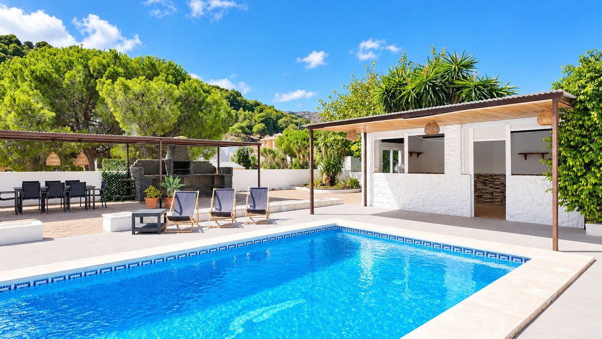 Outdoor swimming pool area with covered bar, dining table, BBQ, lounge chairs, and lush greenery under a clear blue sky.
